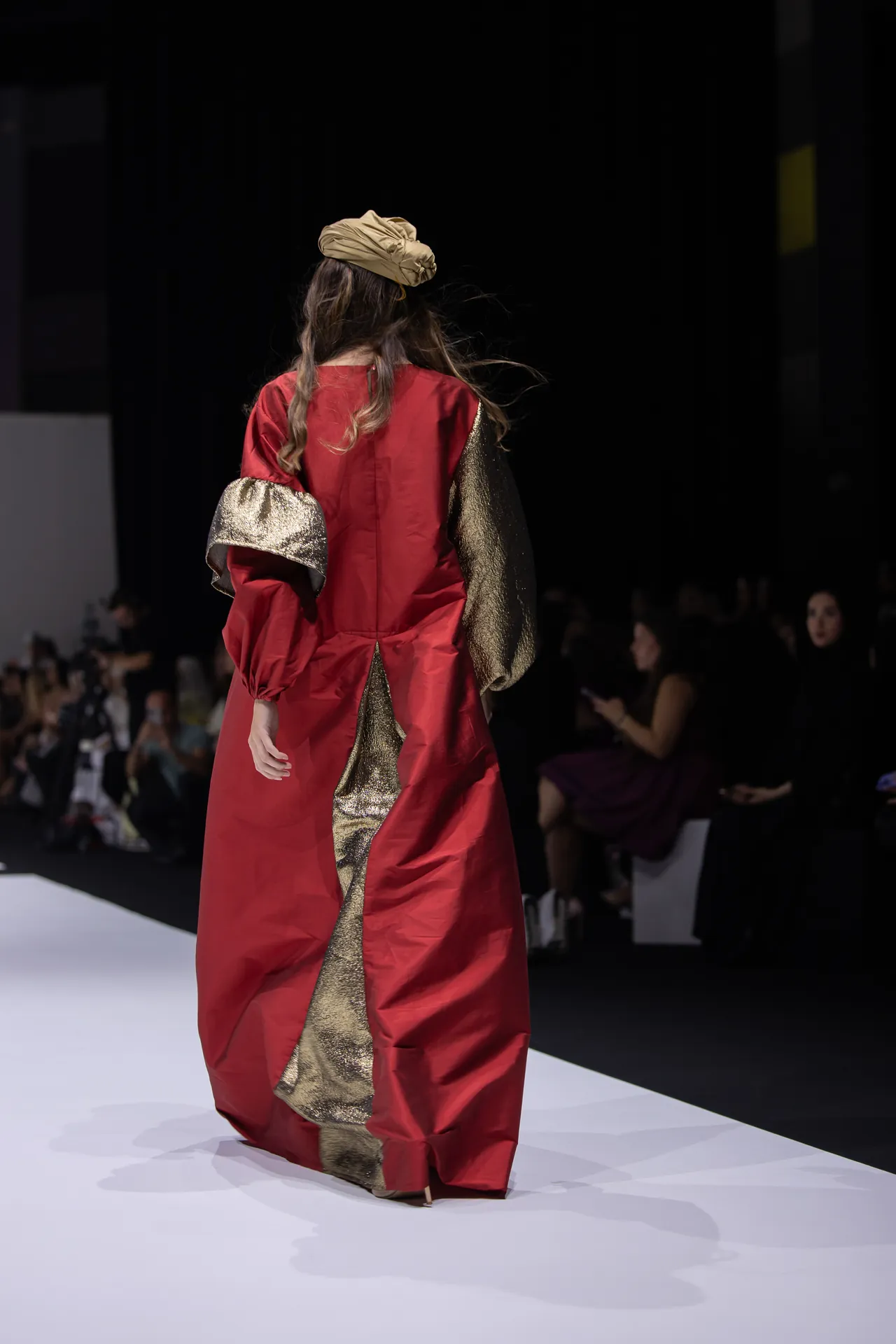 Model with back to camera wearing a red gown with gold metallic panels on a fashion runway; beige head wrap, audience in background.
