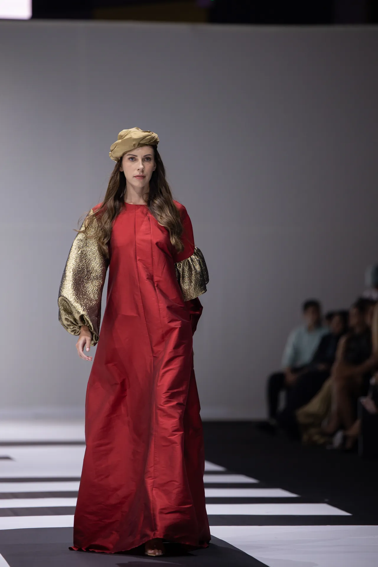 Model walking the runway in a red floor-length gown with metallic gold puffed sleeves and a tan turban-style headpiece.