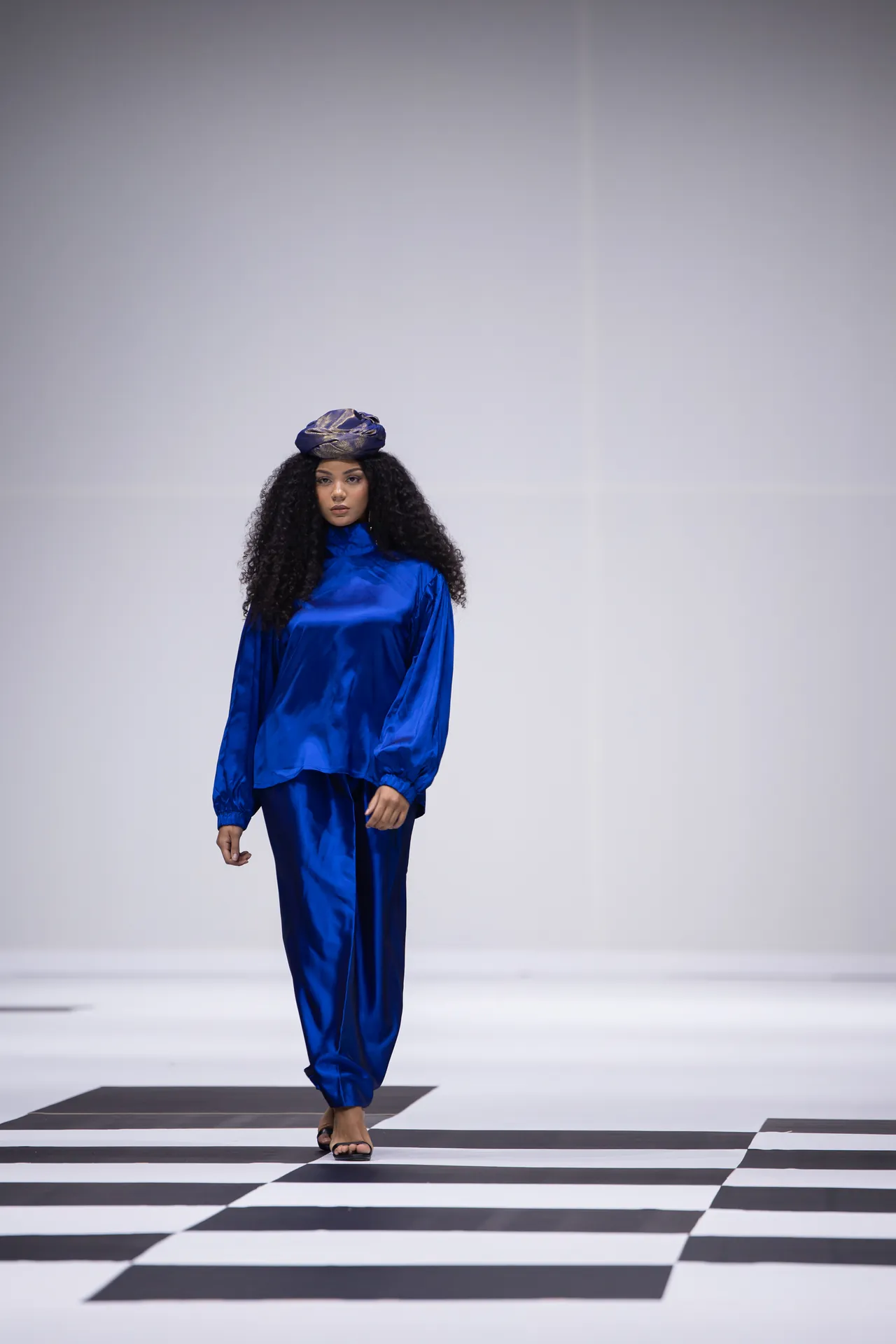 Model walking a fashion runway in a cobalt blue satin outfit with a matching headwrap.
