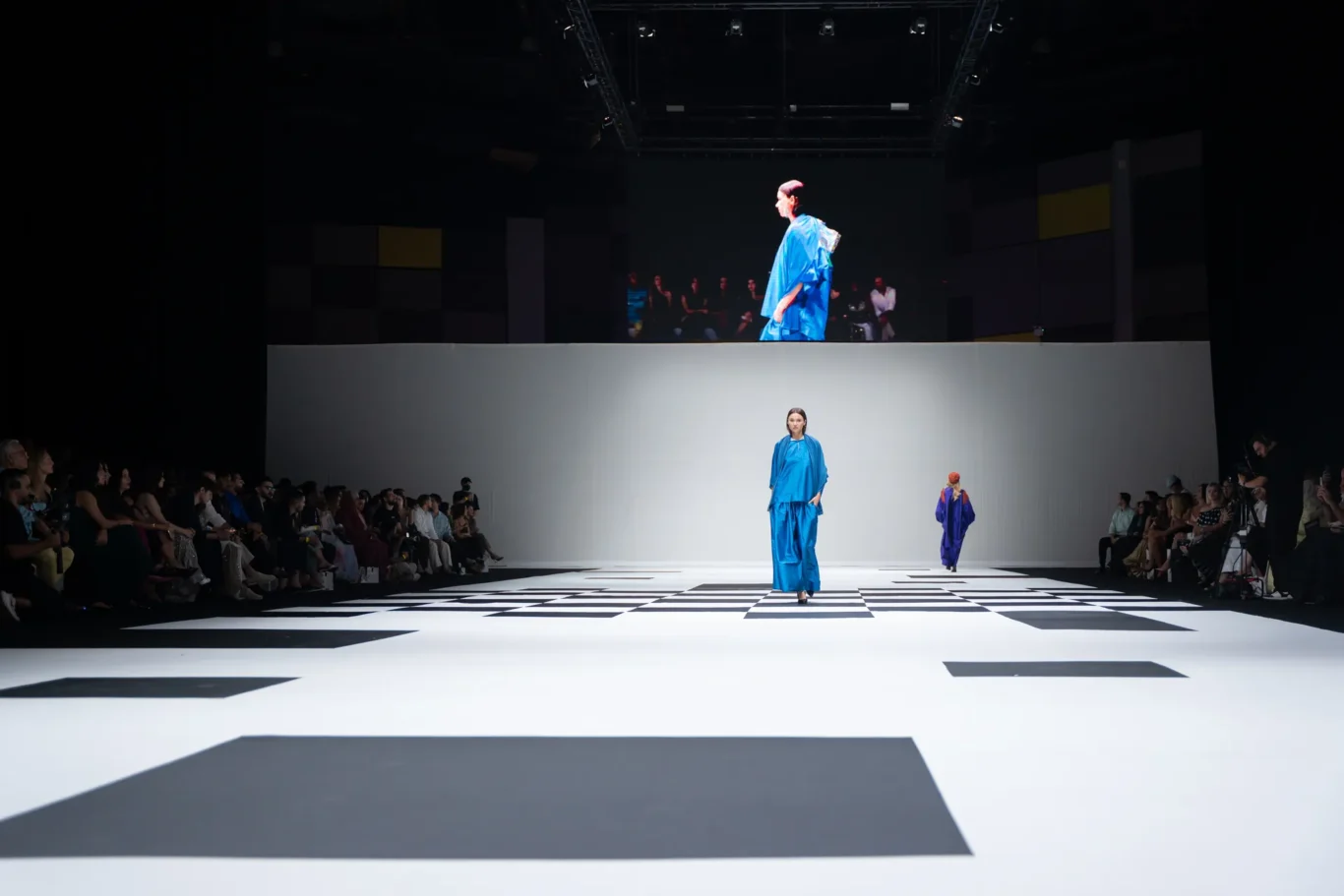 Runway fashion show with models in bright blue outfits walking a black-and-white geometric runway while an audience watches from both sides.