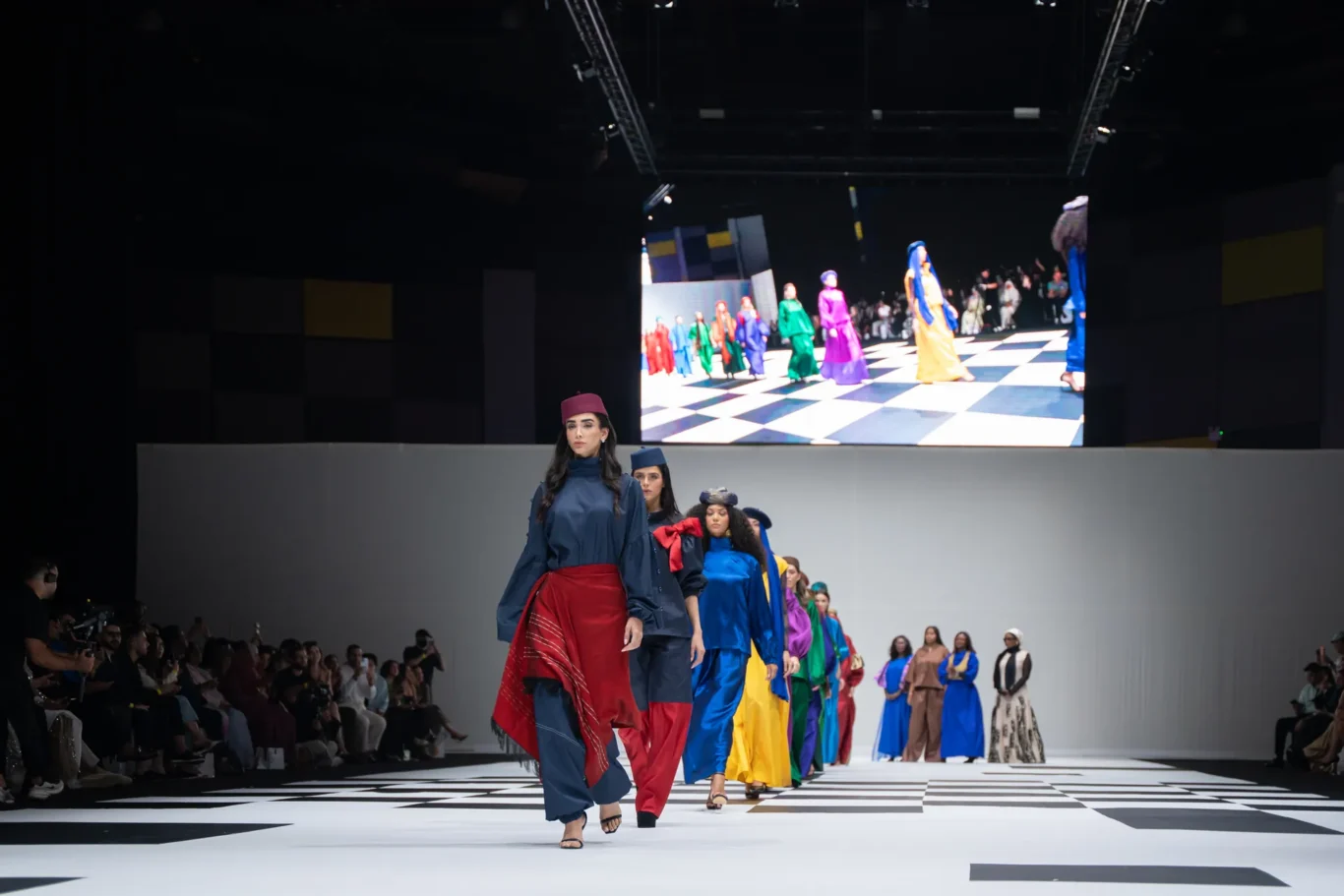 Models walk a fashion runway in bright, multicolored outfits as a large screen watches behind them.