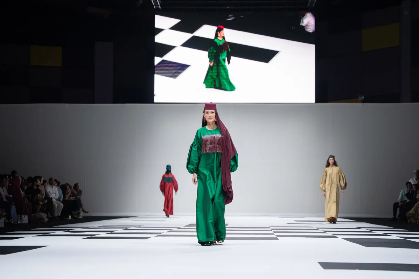 Model walks runway in green outfit with maroon scarf, checkered stage floor, audience on sides, large screen behind.