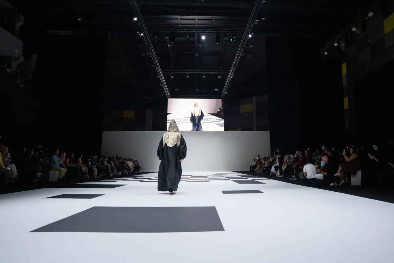 A fashion runway with a model wearing a dark cloaked outfit walking toward a bright screen at the end of a black-and-white checkerboard runway, surrounded by seated audiences on both sides.