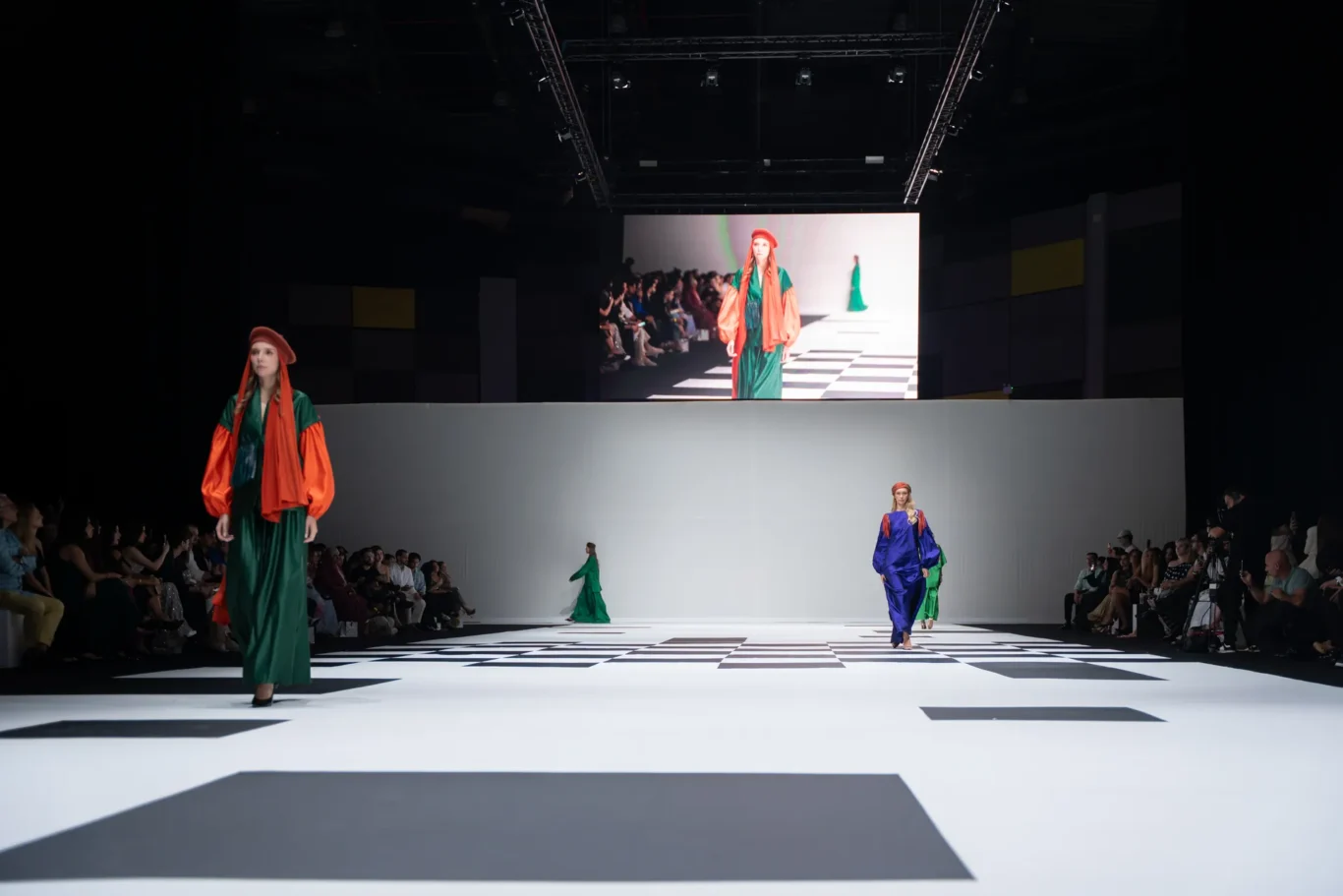 Models in colorful, oversized robes walk a black-and-white checkered runway as a seated audience watches from both sides.