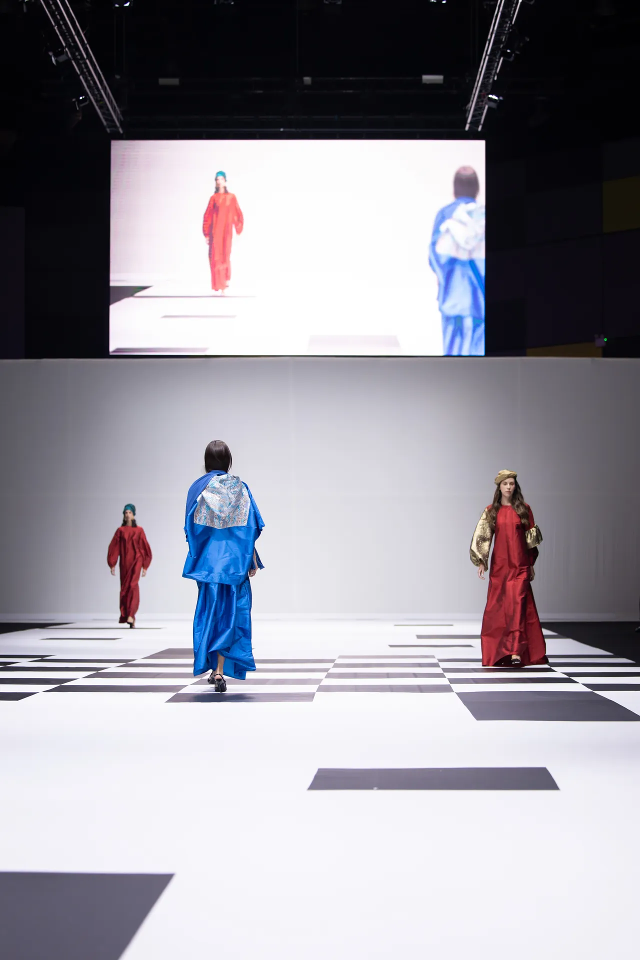 Models walk a white runway in bold blue and red outfits, with a large screen above and a black-and-white checkered floor pattern below.