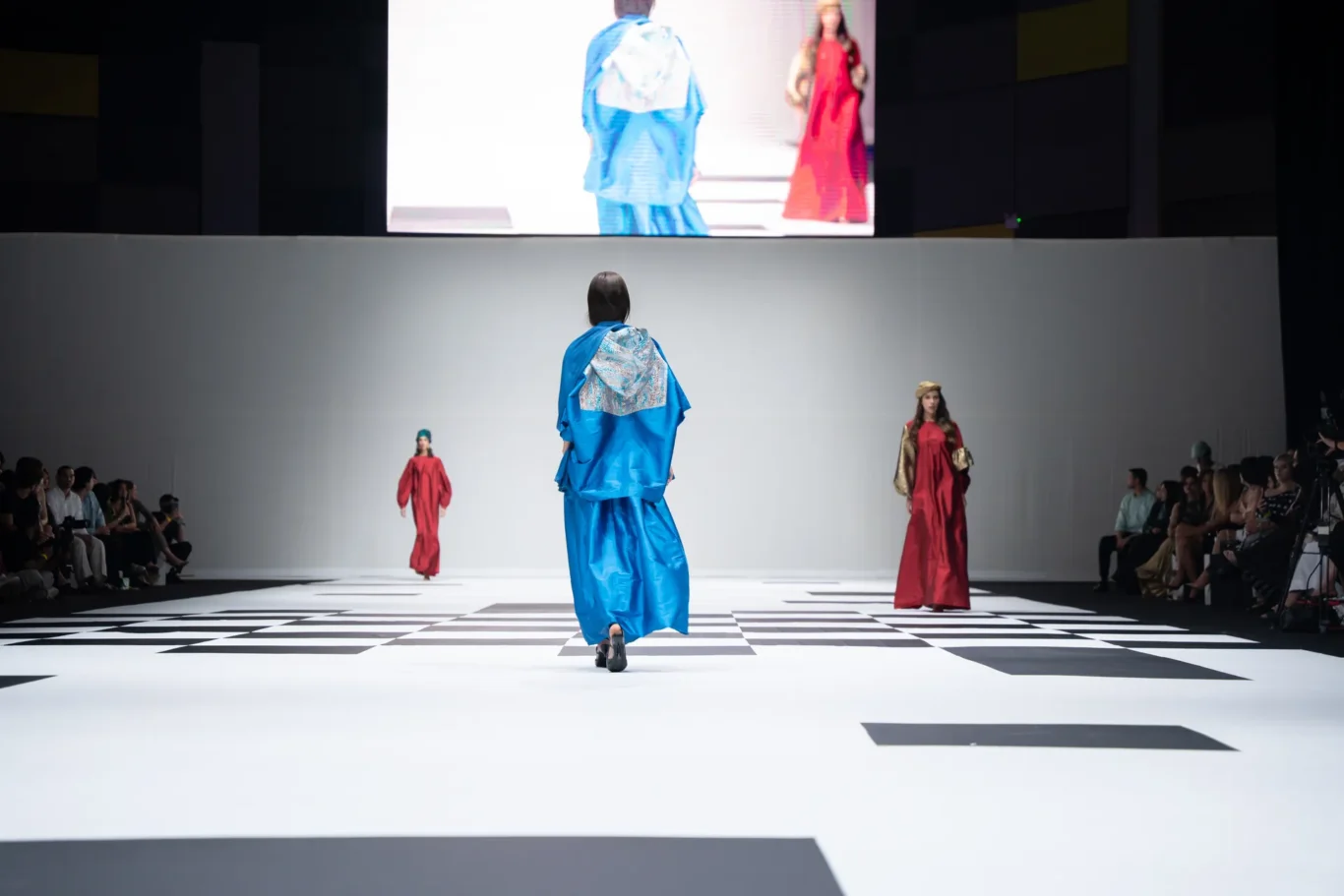 Model walks away from camera on a white runway with black geometric patterns; wearing a bright blue flowing dress as others in red outfits stand on the sides and a large screen looms above.
