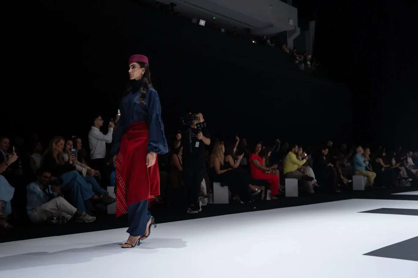 Model walking a runway in a navy outfit with a red sash and magenta hat; audience and photographers along darkened venue visible in the background
