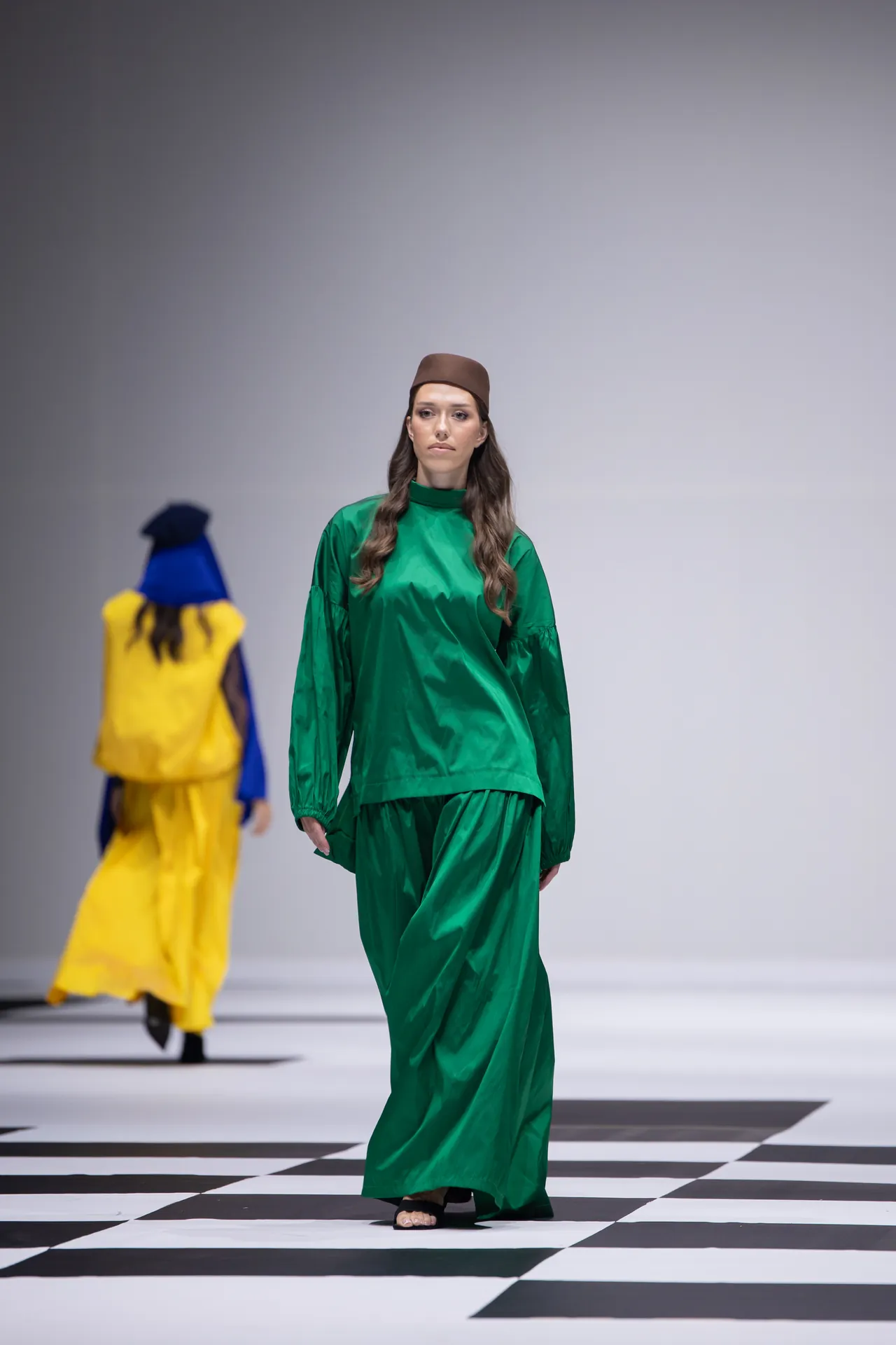 Model walks a fashion runway in a green satin outfit and brown cap, on a black-and-white checkered floor with another model in yellow in the background.