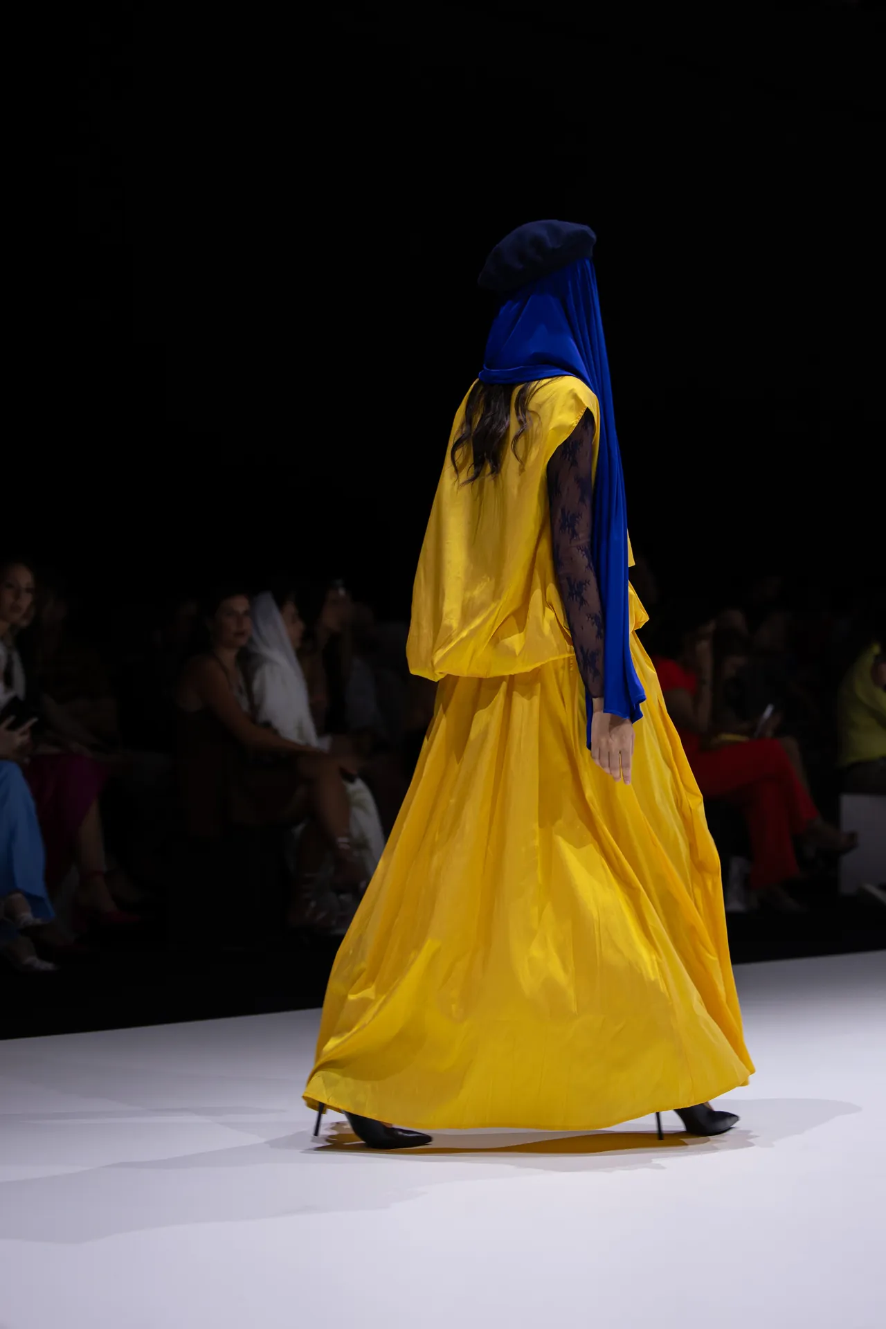 Back view of a model walking a runway in a bright yellow gown with a blue headscarf, audience in darkness behind her.