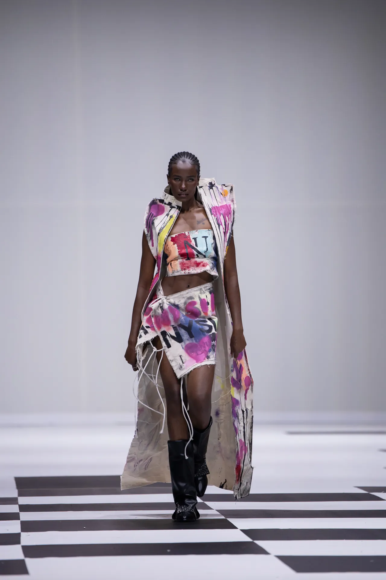 Model walking a runway in a paint-splattered, color-rich outfit against a plain backdrop on a black-and-white checkered floor.
