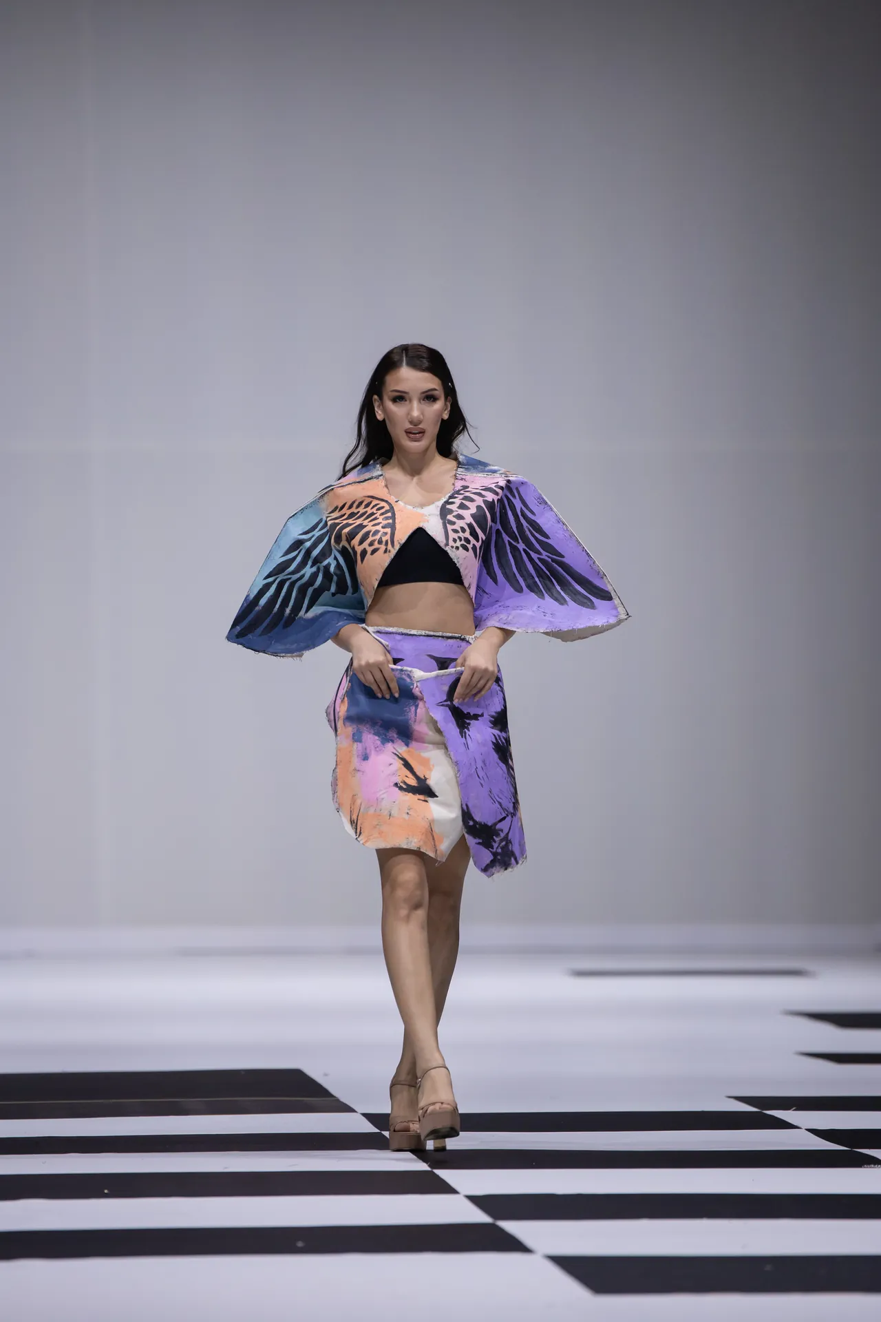 Female model walks a fashion runway in a colorful abstract-print two-piece outfit with cape-like sleeves and platform sandals.