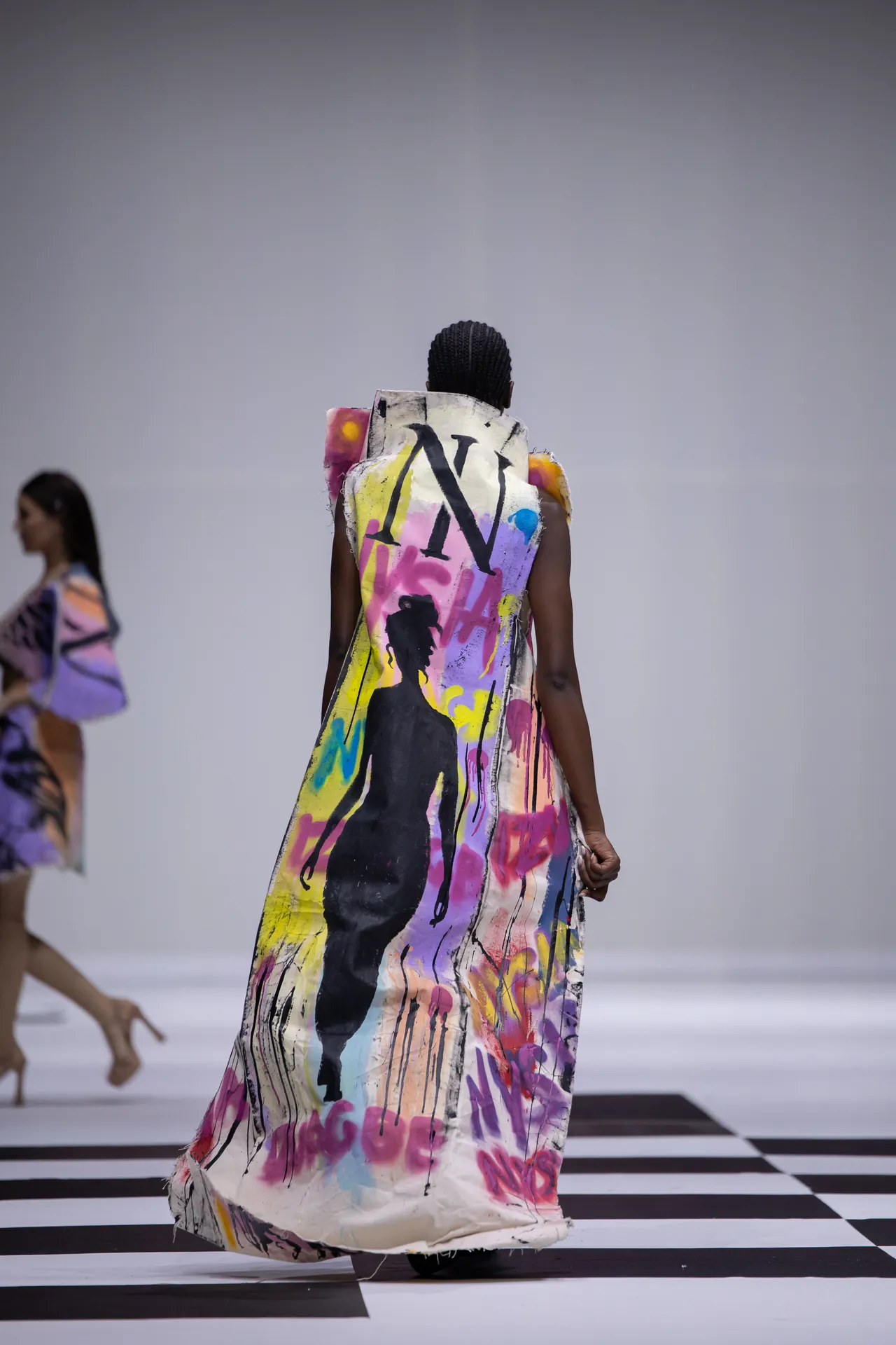 Back view of a model wearing a long graffiti-print dress with a bold multicolor pattern, walking away on a black-and-white checker runway.