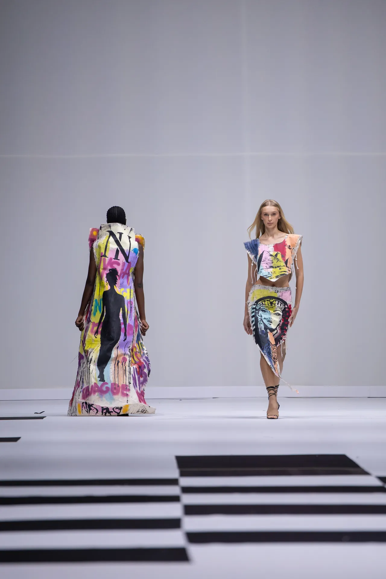 Two models walk a runway in graffiti‑print outfits; the front model wears a cropped top and skirt, the back model wears a long colorful dress.