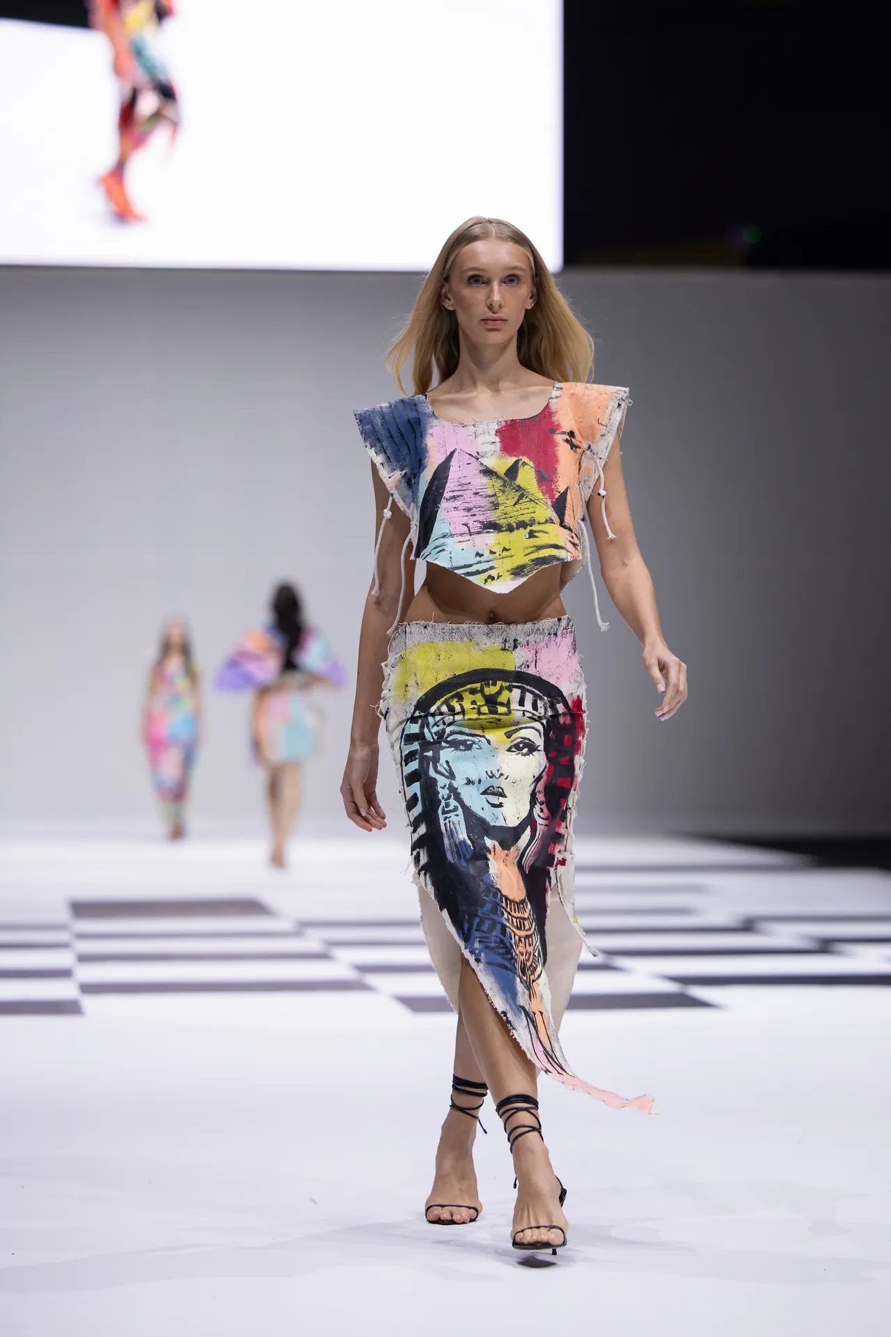 Model walking the runway in a colorful abstract two-piece outfit with fringe and strappy sandals.
