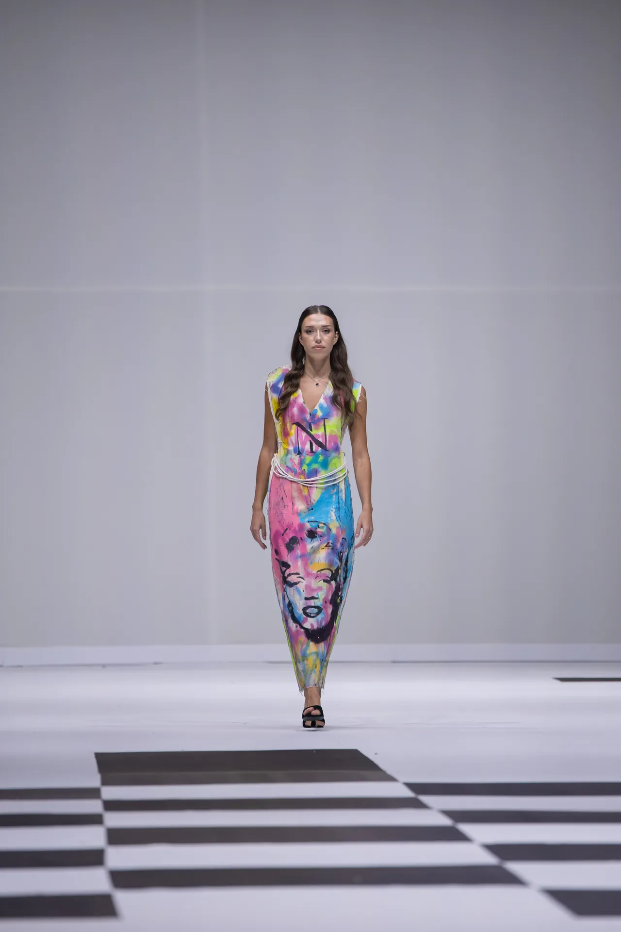 Model walks a white runway wearing a vibrant paint-splatter maxi dress with a portrait print and black sandals.