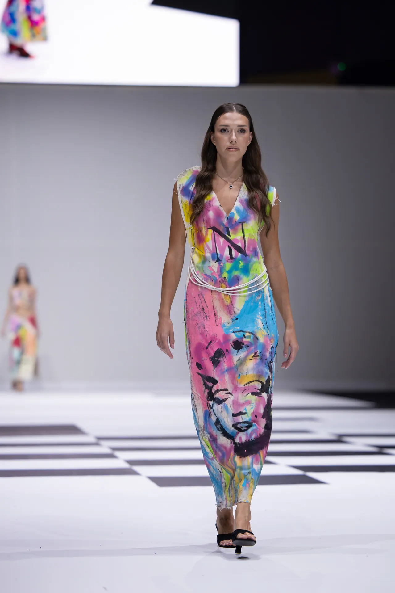 Model walking a fashion show wearing a sleeveless, multicolor abstract print dress with a face design on the front.