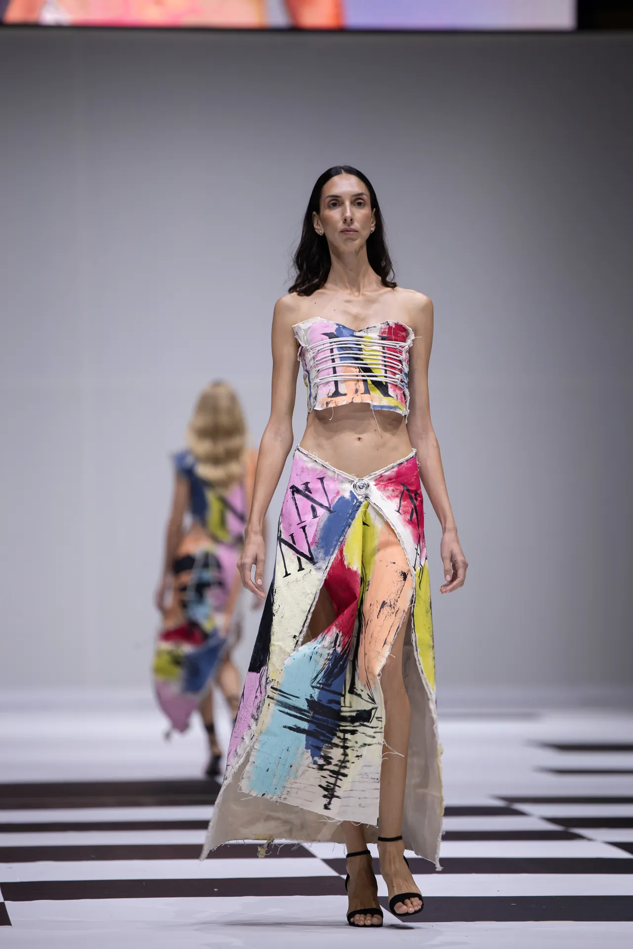 Runway model walks the catwalk in a strapless, multicolored abstract-print crop top and matching long skirt with a high slit.