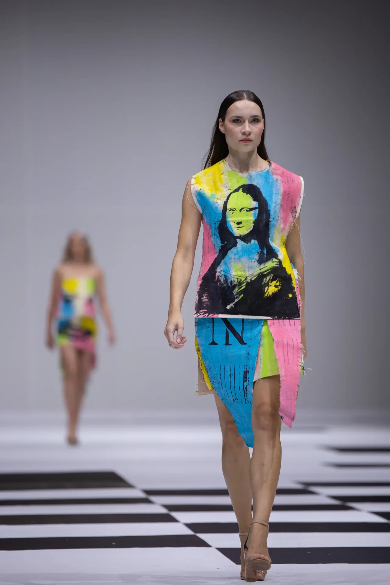 Model walking a runway in a sleeveless, multicolor dress with a Mona Lisa portrait print.