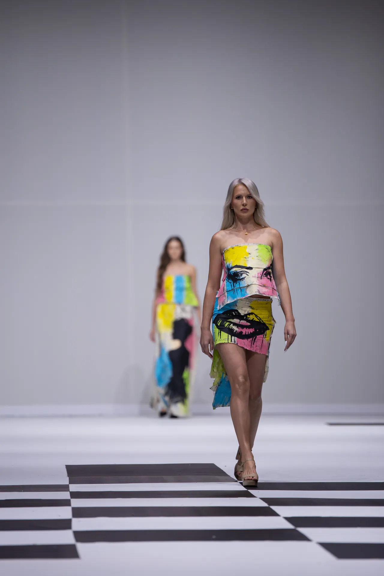 Model walking a fashion show in a strapless, multicolored abstract-print dress on a black-and-white checkered runway.