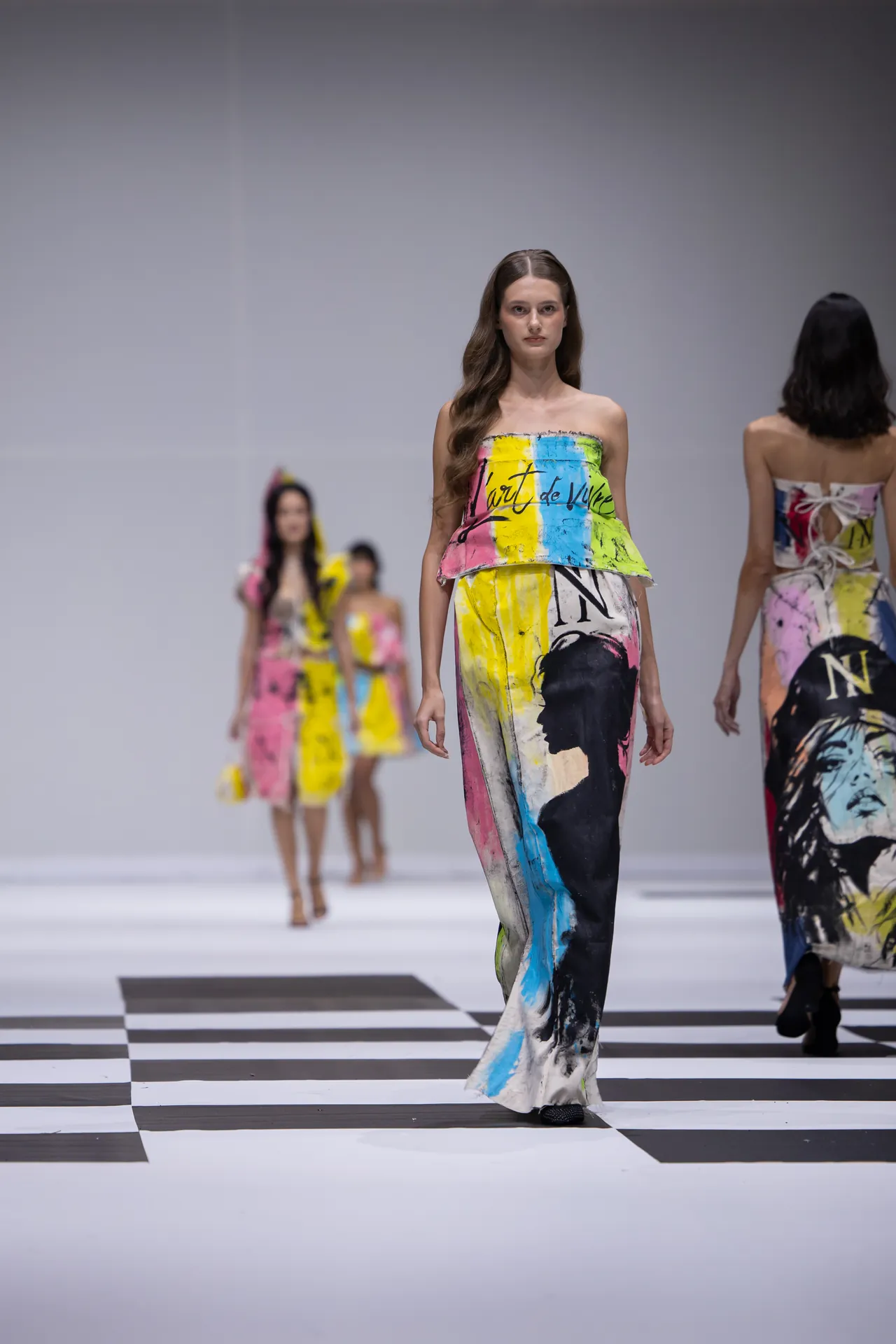 Center model walks a strapless, multicolored abstract-print outfit on a black-and-white checkered runway; others follow in background.