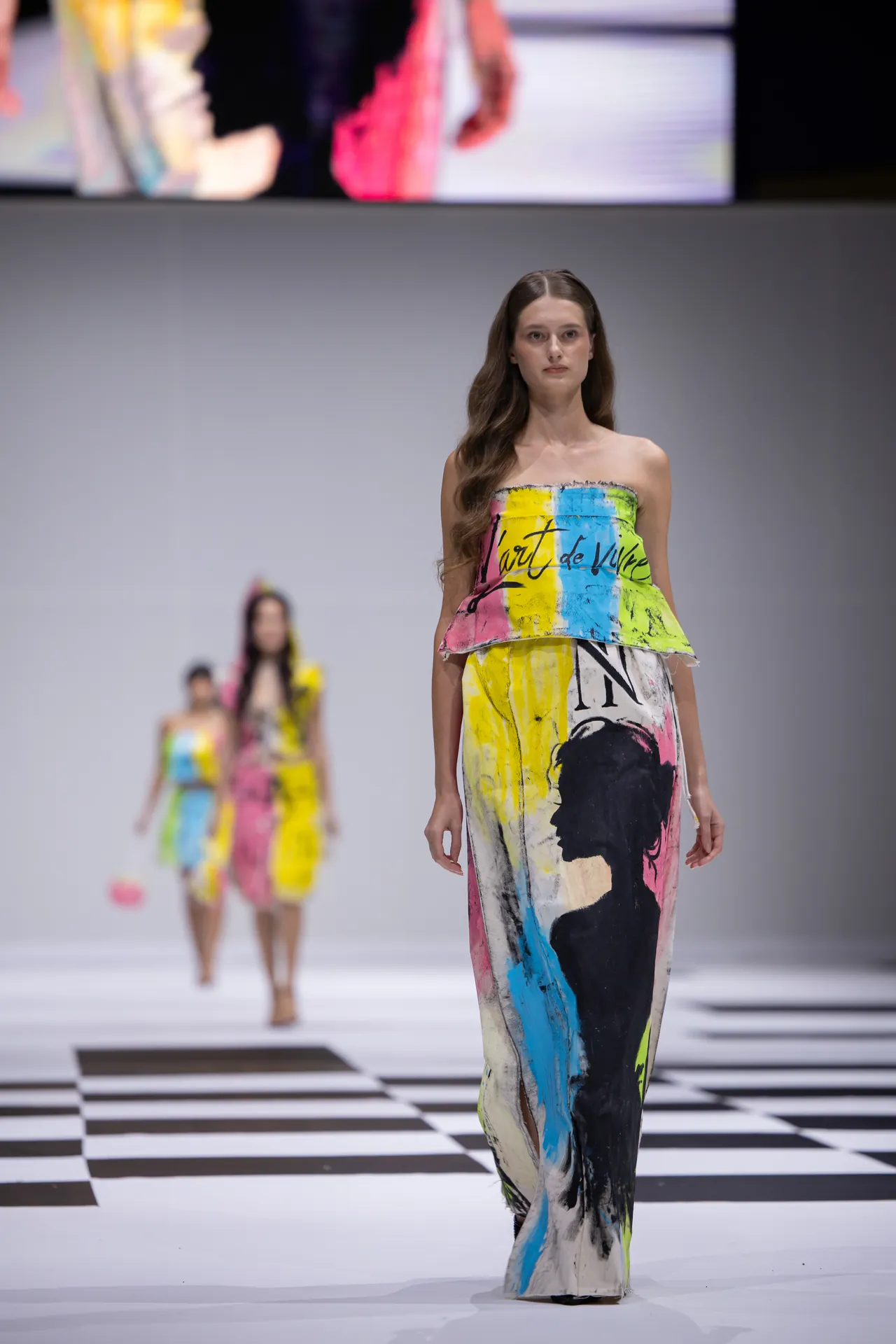 Female model walks a runway in a strapless, abstract-colorful gown with black, pink, blue, and yellow brushstroke patterns; checkered floor backdrop.