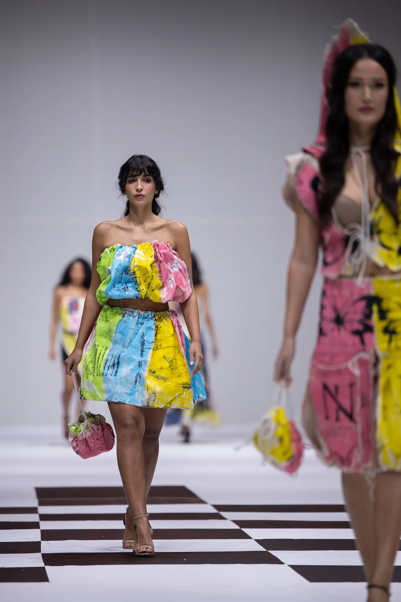 Model walks a runway in a bright multicolor strapless crop top and matching skirt, carrying a pink polka-dot bag.