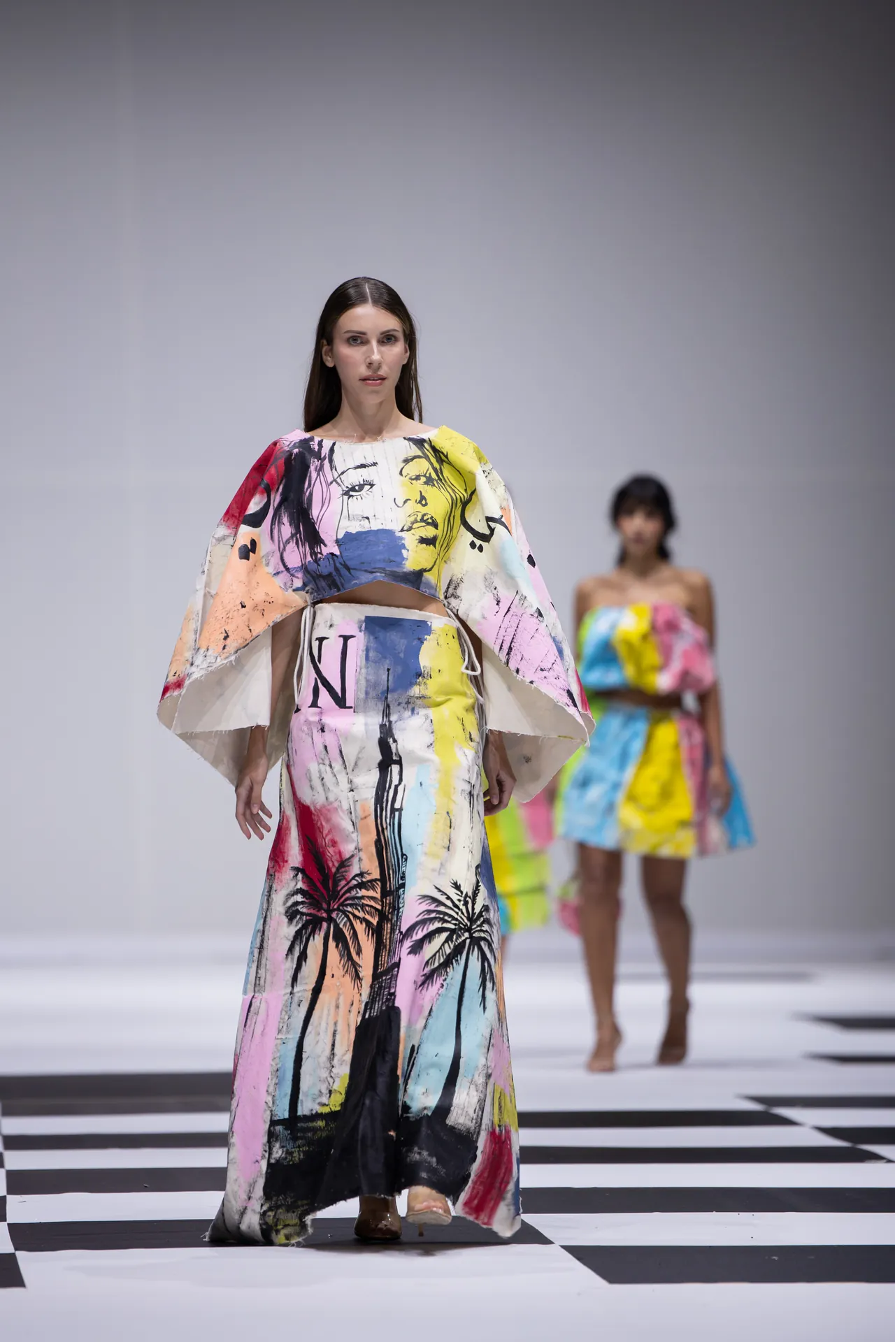 Model walks the runway in a long, abstract-print multicolor dress with cape-like sleeves.