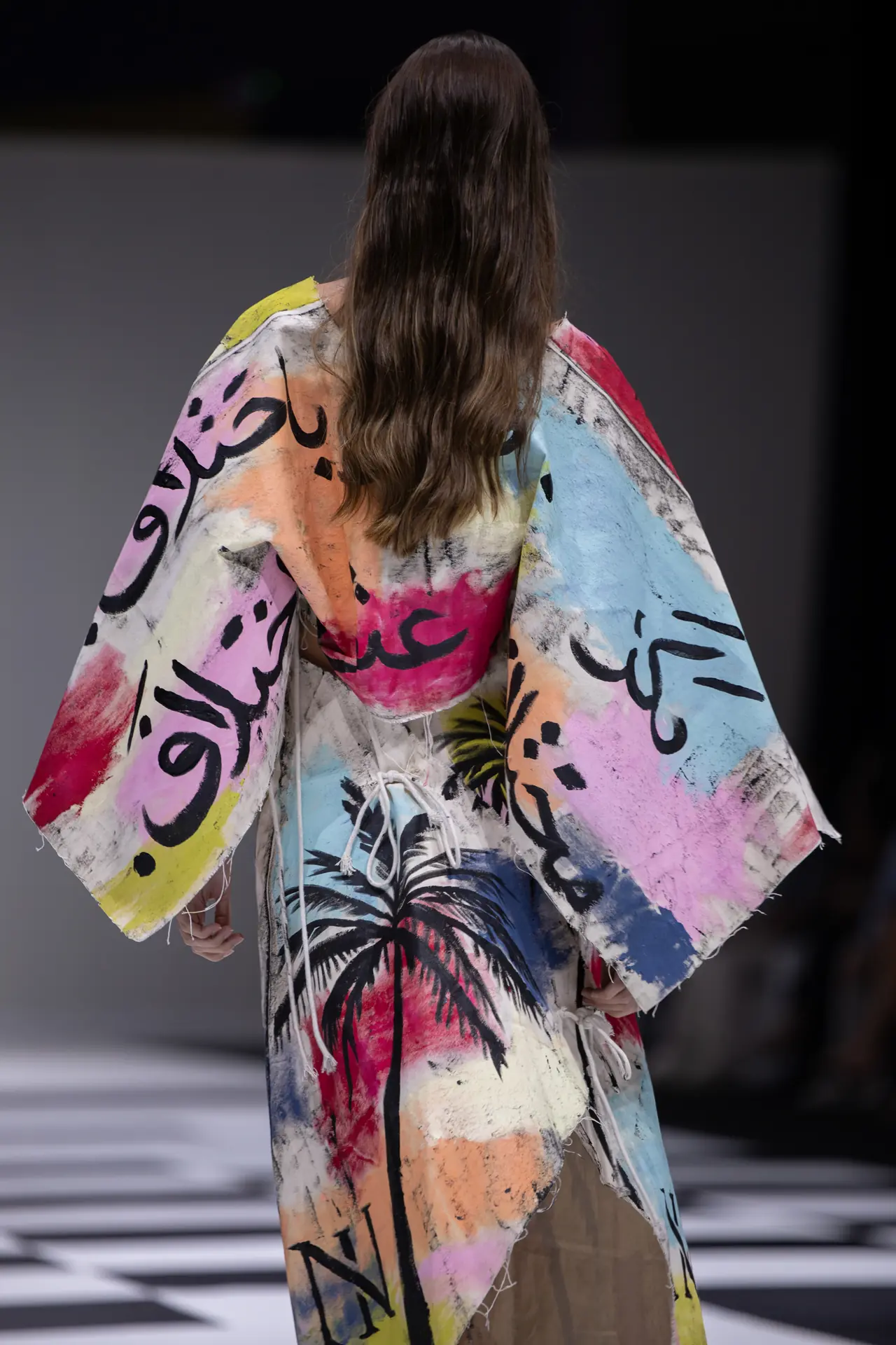 Model walking away on a fashion runway wearing a colorful abstract-painted robe with fringe, seen from behind against a checkered floor backdrop.