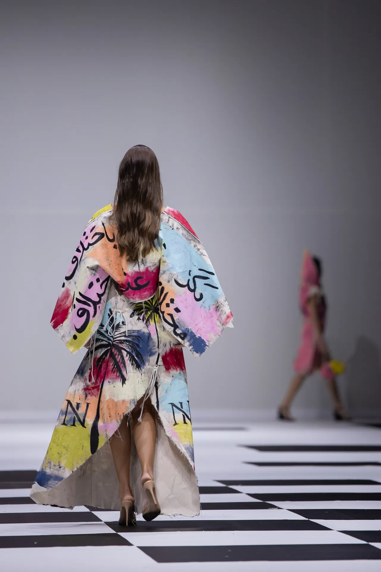 Model walking away on a black‑and‑white checkered runway, wearing a colorful abstract cape, back view