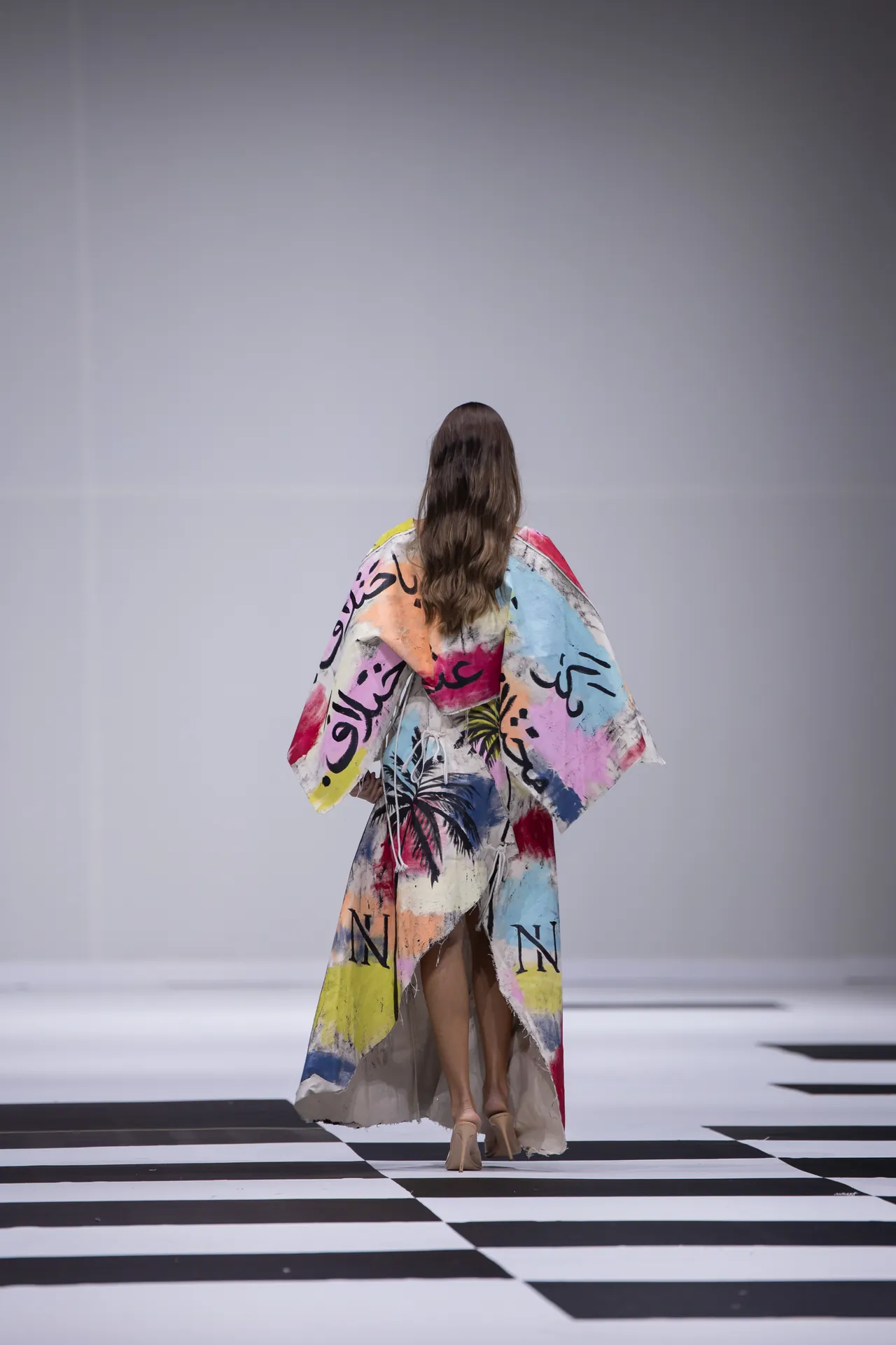 Person walking away on a fashion runway, wearing a flowing colorful cape with graffiti-style patterns over a black-and-white checkered floor.