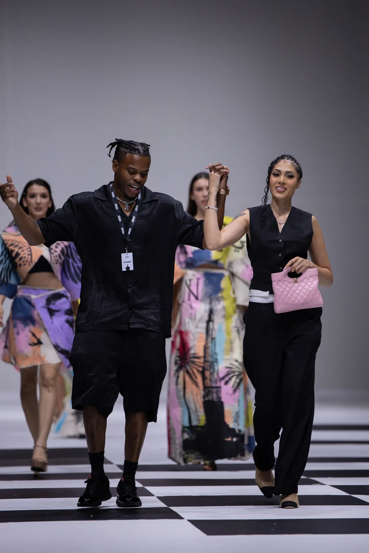 Two models walk hand-in-hand on a black-and-white checkered runway during a fashion show, with others in colorful outfits in the background.
