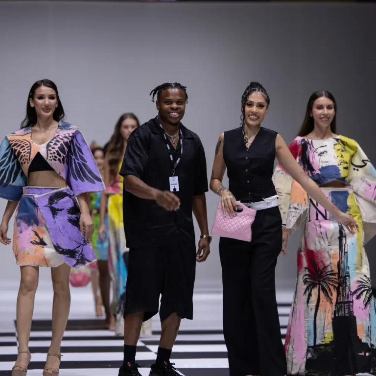 Group of models walking a fashion-show runway; vibrant printed outfits and a model in black with a pink quilted bag lead the line.