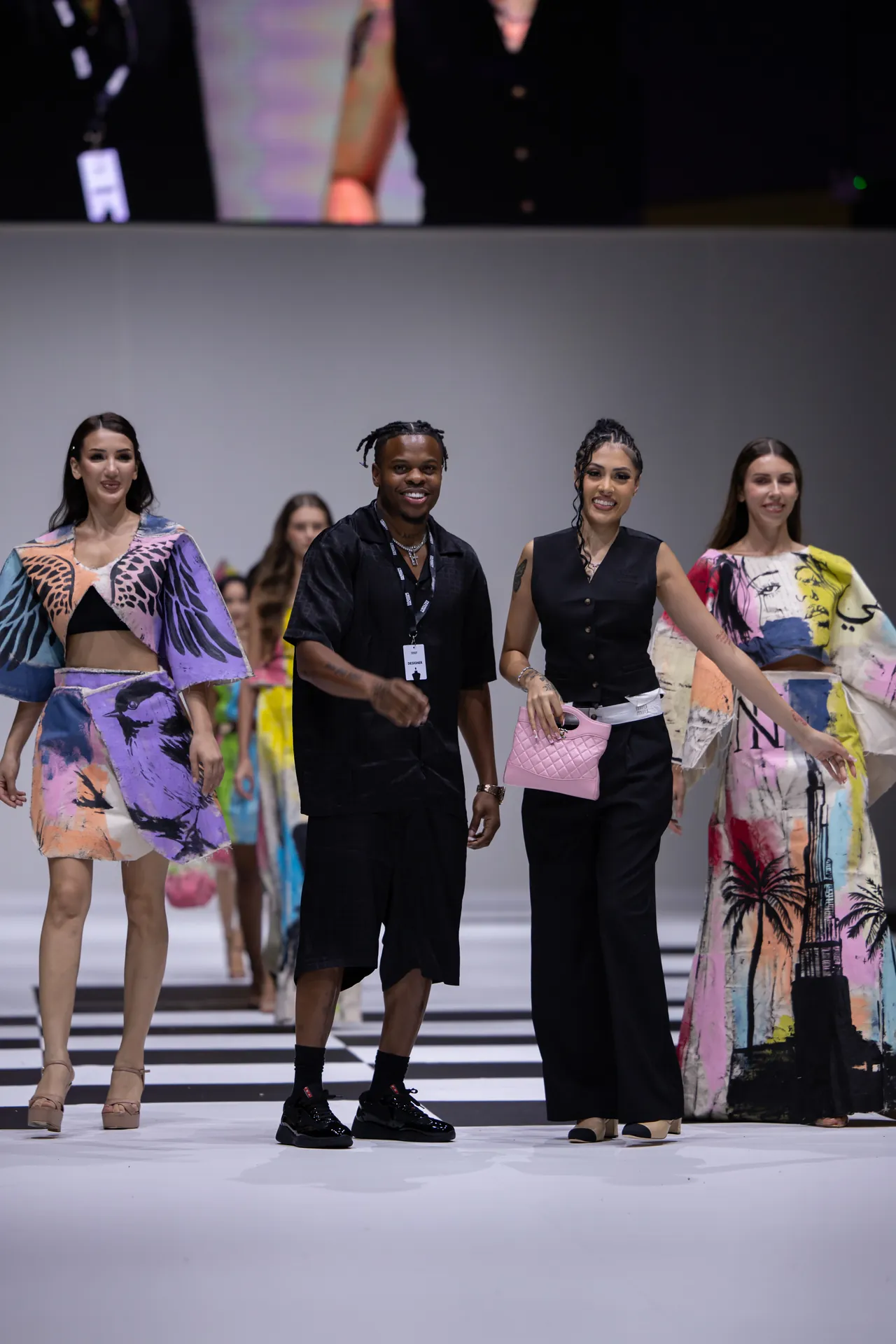 Group of models walking a fashion-show runway; vibrant printed outfits and a model in black with a pink quilted bag lead the line.