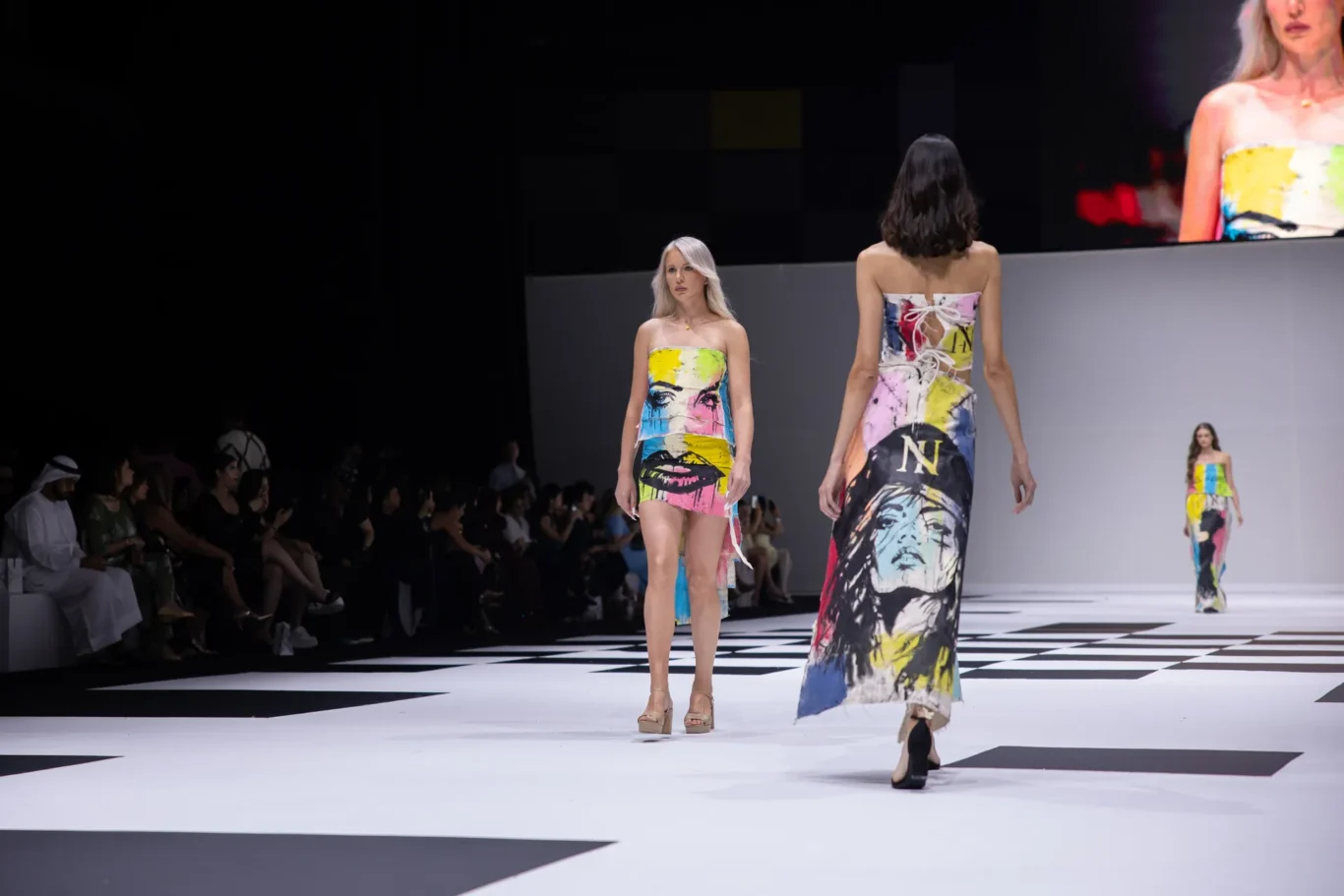 Model in a bright, strapless graphic dress walks the runway as an audience watches from the left.