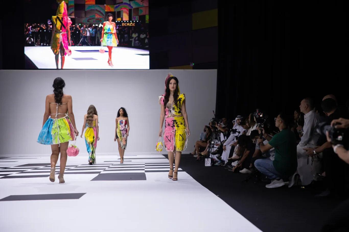 Models in bright, multicolor outfits walk a fashion runway as photographers and an audience observe from the right.