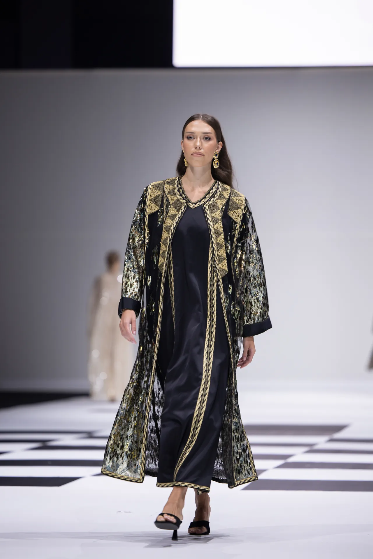Female model walks a runway in a black and gold embellished robe with geometric trim.