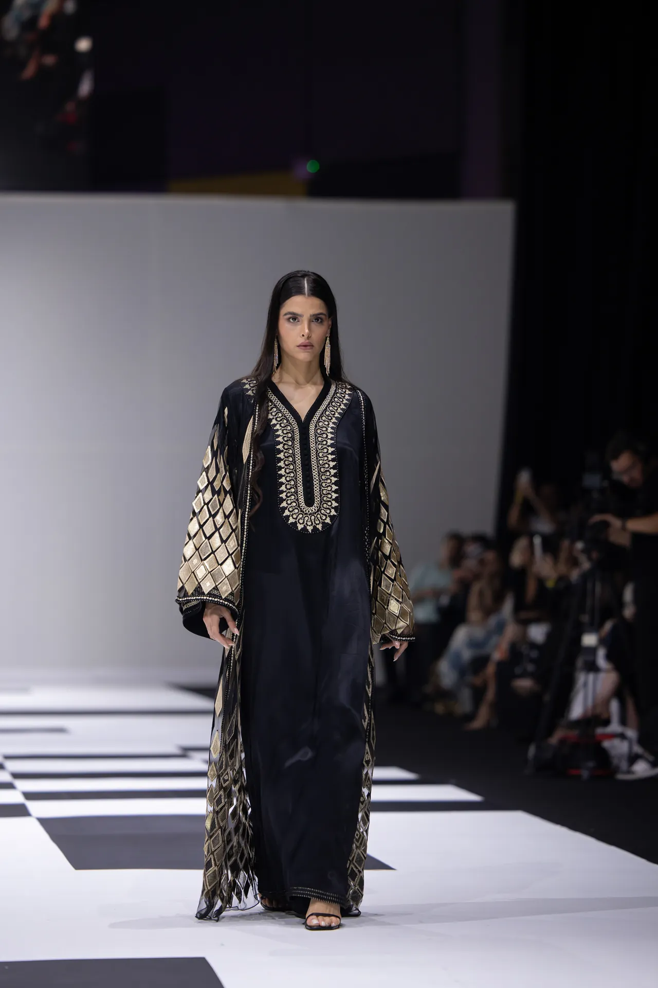 Model on a runway wearing a black, gold-embroidered gown; audience blurred in the background on the sides of the catwalk.