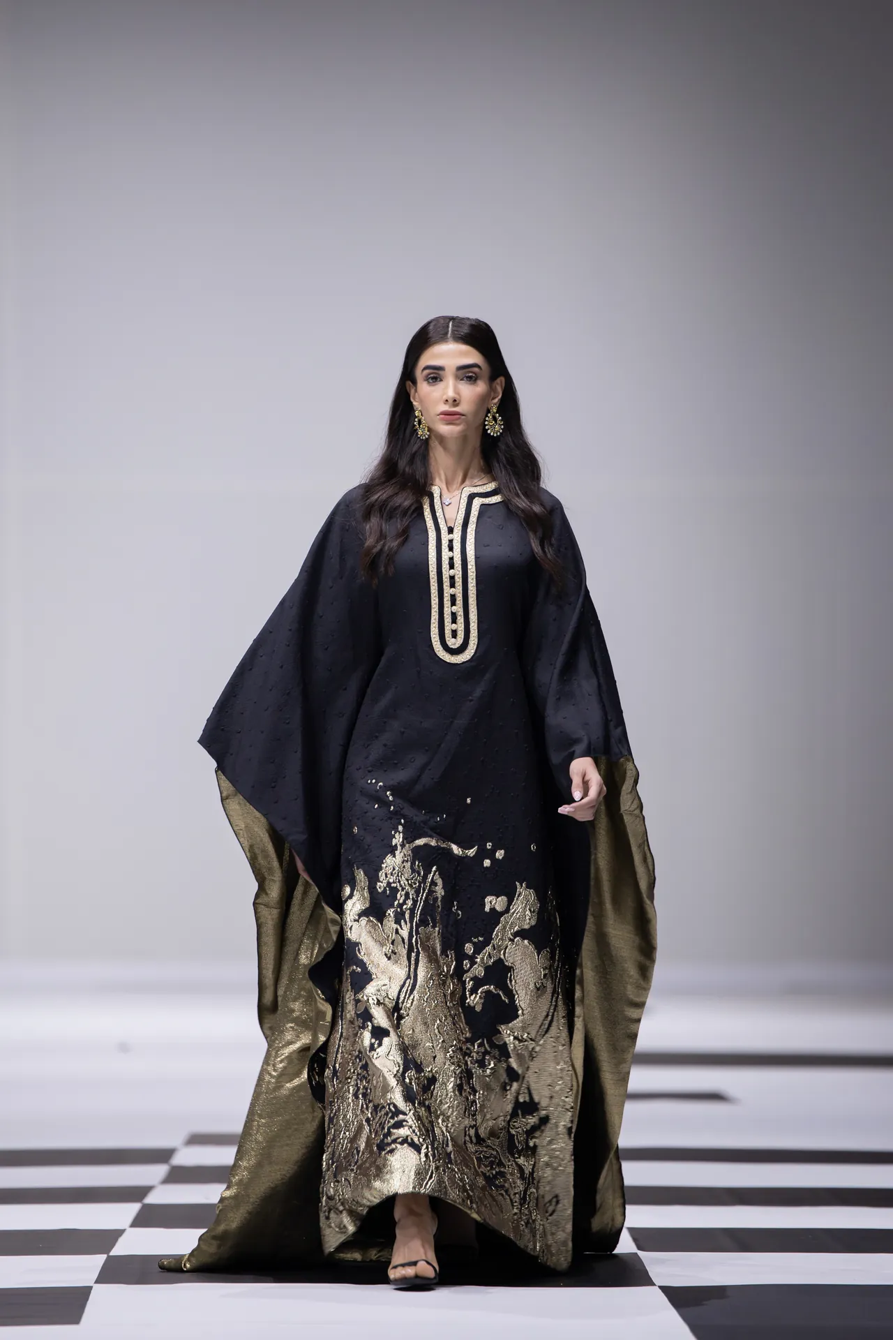 Model walking the runway in a black, gold-embroidered kaftan with a gold-trim neckline and wide sleeves, on a black-and-white checkered floor.