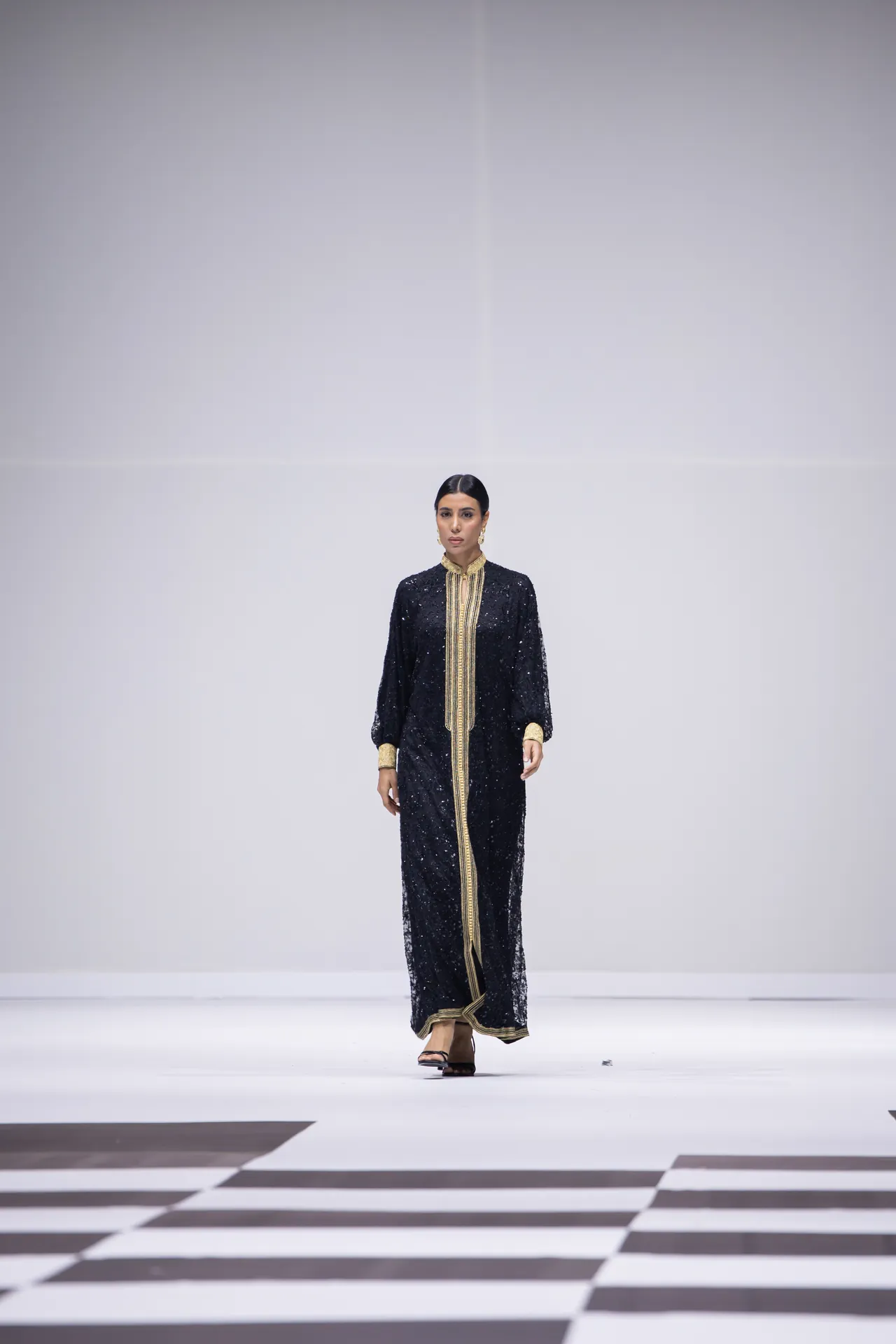 Model walking a white runway in a floor-length black sequin dress with gold trim and cuffs.