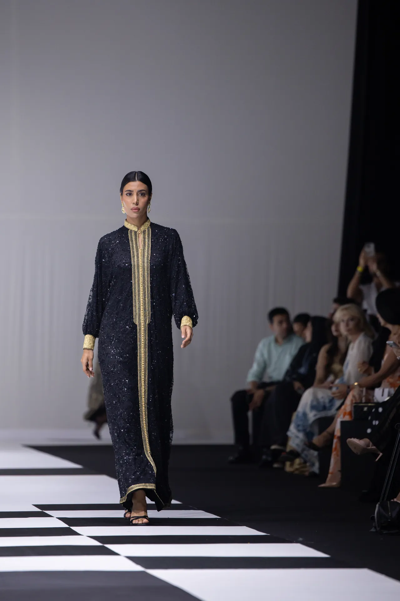 Model walking a runway in a black sequin gown with gold vertical trim and cuffs, audience watching on the right.