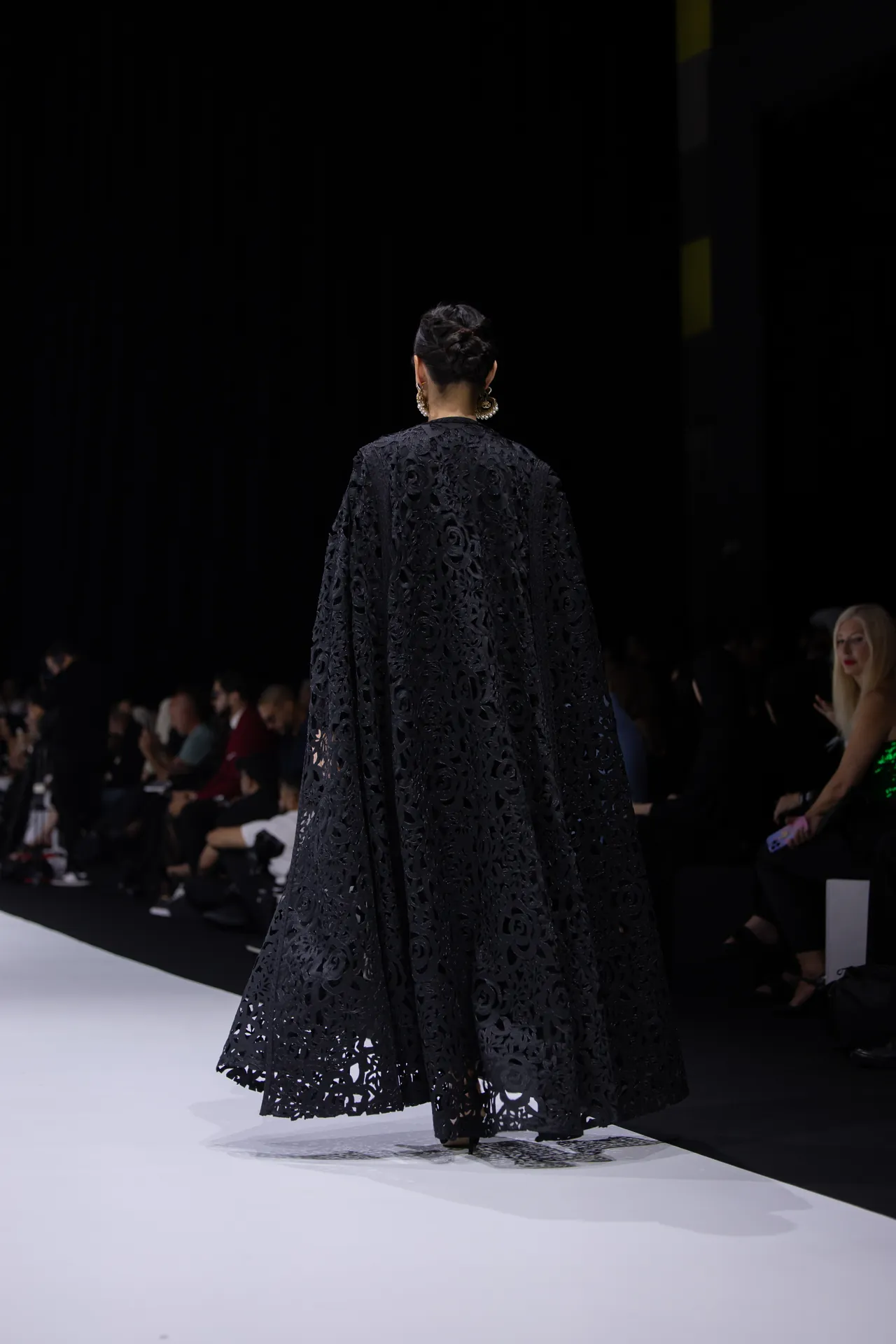 Model walking away on a fashion runway in a long black lace cape, ornate earrings visible, audience in background.