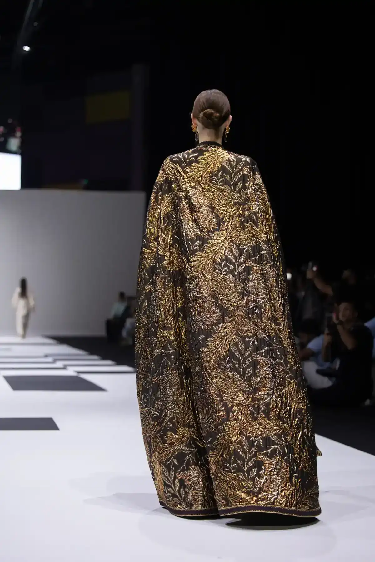 Model walking runway in a dark, gold-embroidered cape with floral patterns; hair in a bun, audience in background.