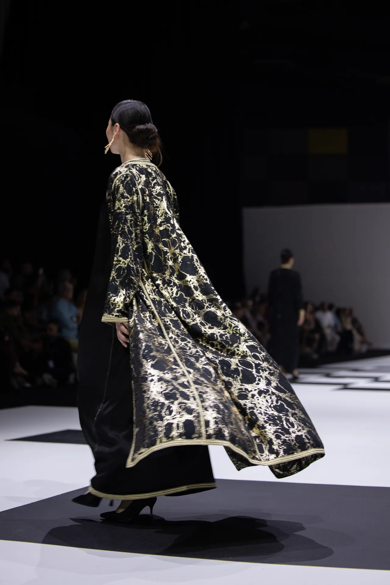 Model walking the runway in a black and gold brocade cape with intricate floral patterns and gold trim, audience blurred in the background.