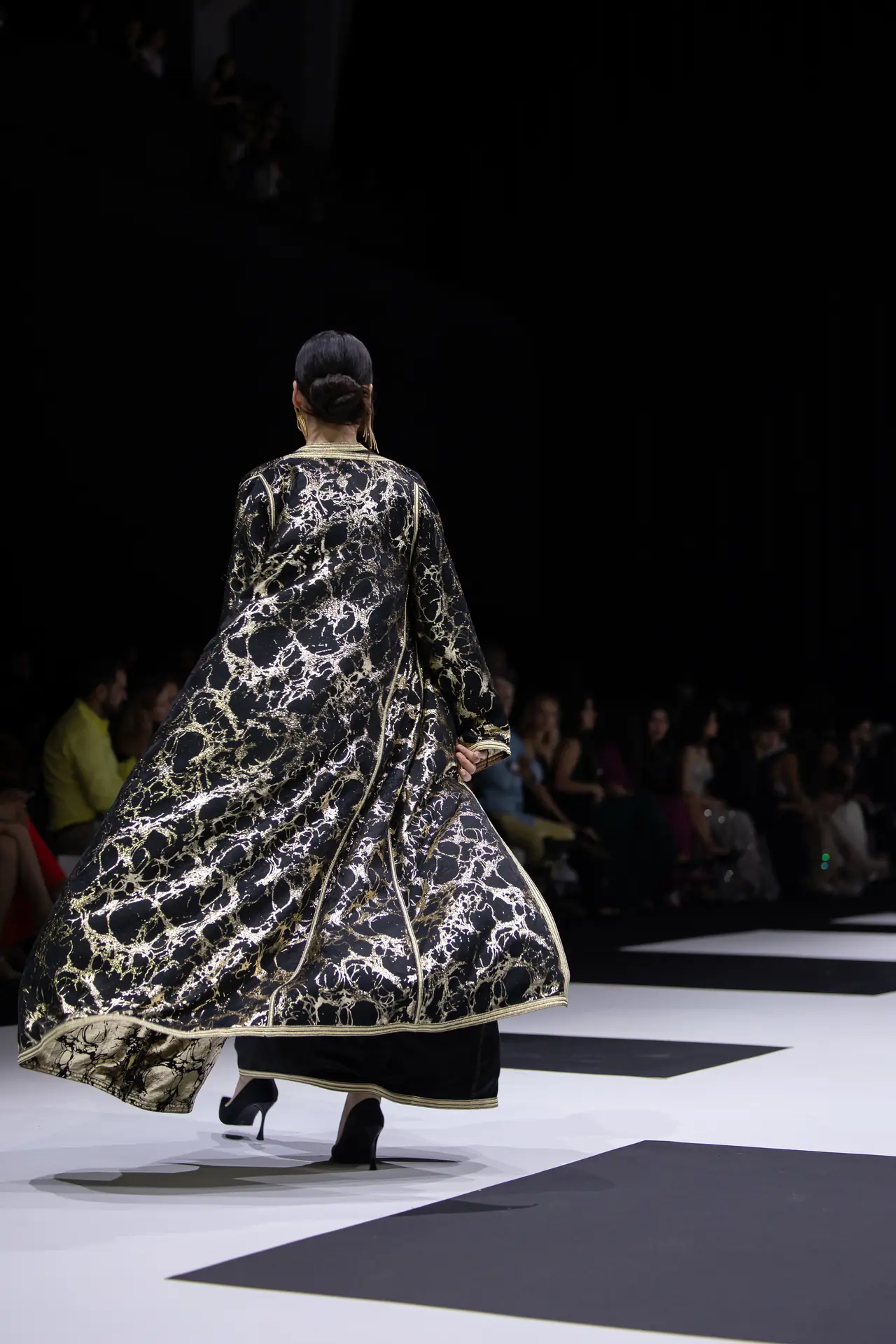Model walking away on a runway in a black and gold marbled cape-like garment; audience in the dark background.