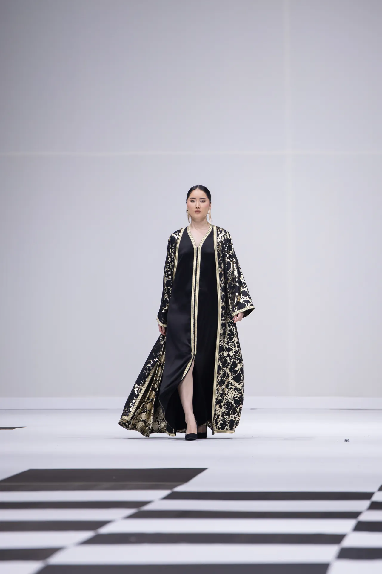 Model walking a white runway in a black and gold embroidered robe with wide sleeves and a slit in the front.