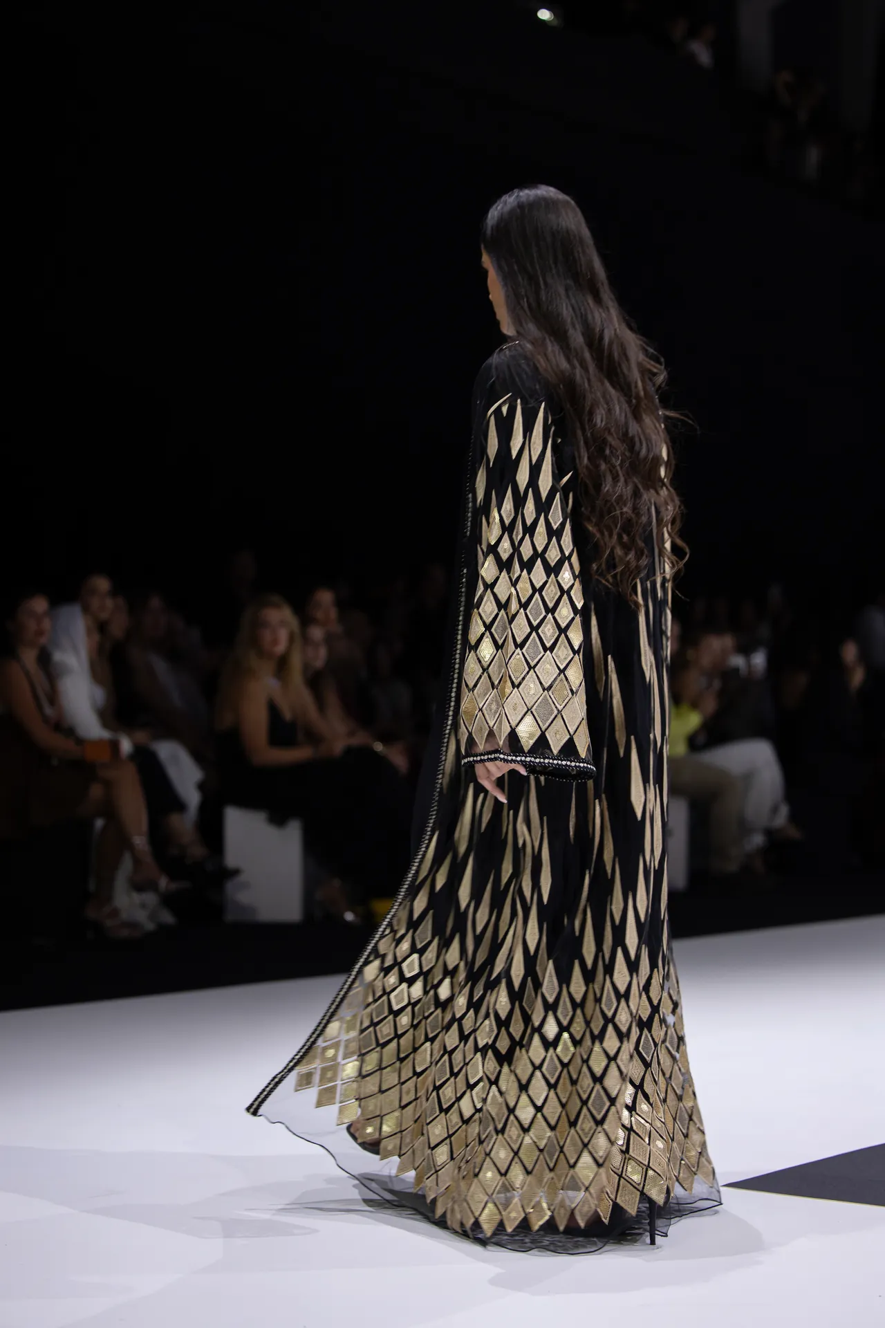 Model walks the runway in a black-and-gold diamond-pattern gown, with a blurred audience in the dark background behind her.