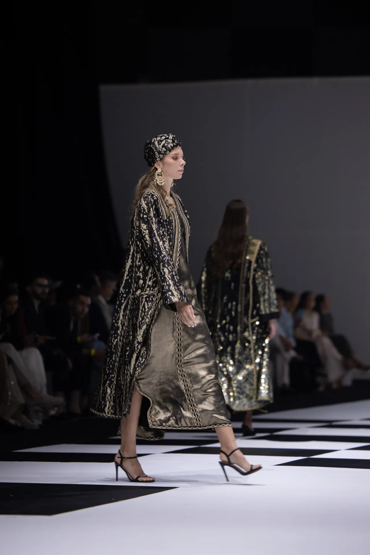 Model walking the runway in a black-and-gold metallic ensemble with a matching head wrap and large gold earrings (side profile).