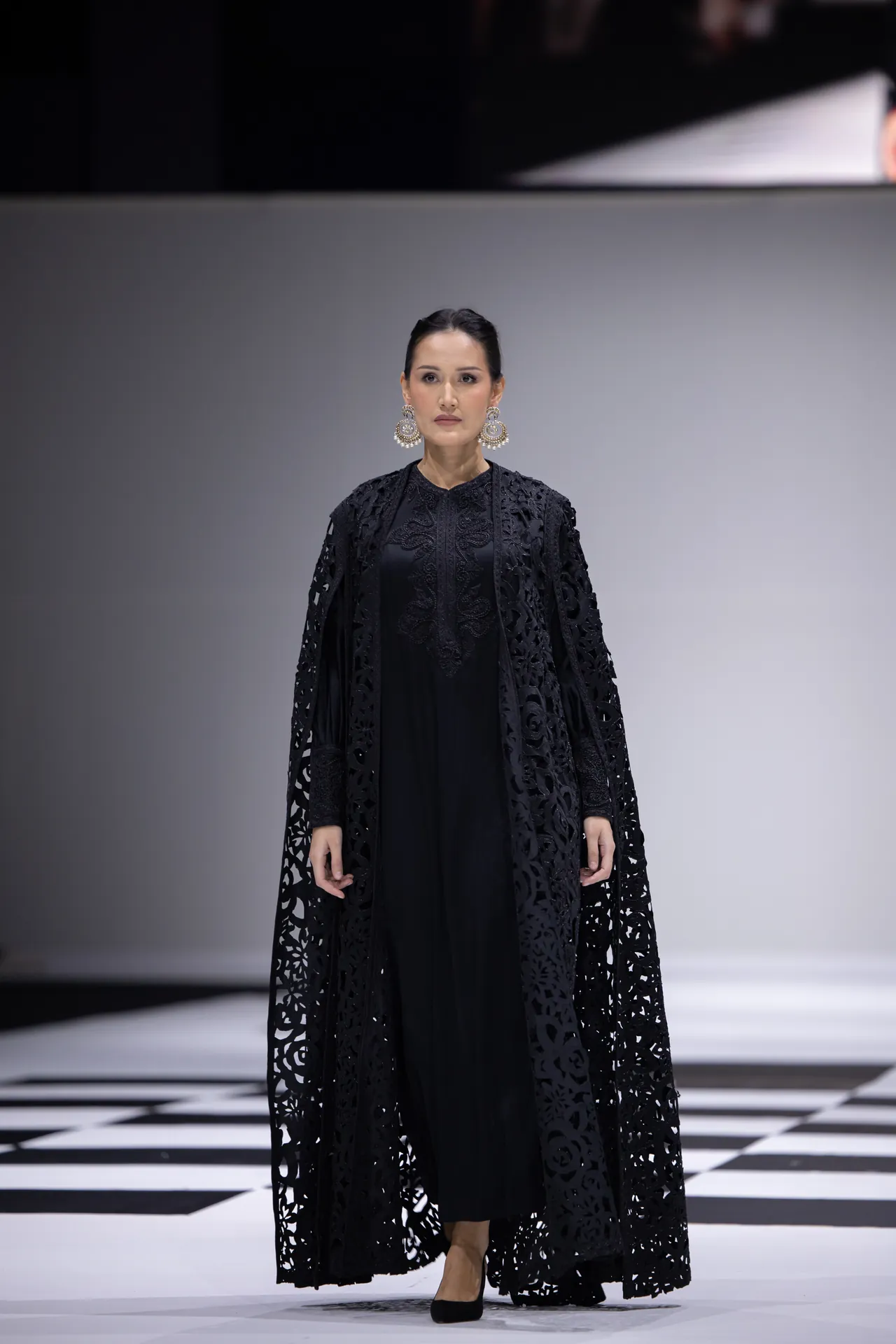 Model walking a fashion show in a black lace cape over a black dress, wearing large circular earrings against a checkered runway backdrop.