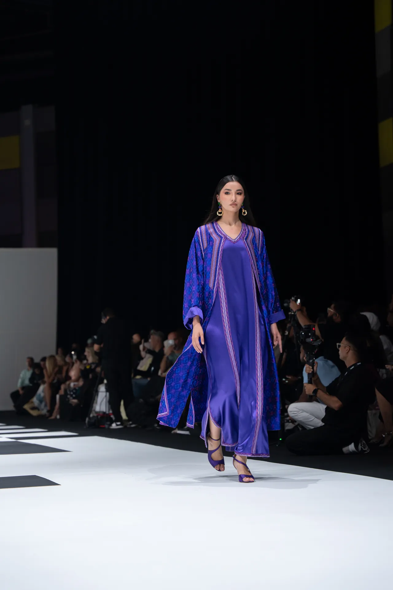 Female model walks the runway in a purple patterned kaftan over a satin purple dress, with photographers in the background.