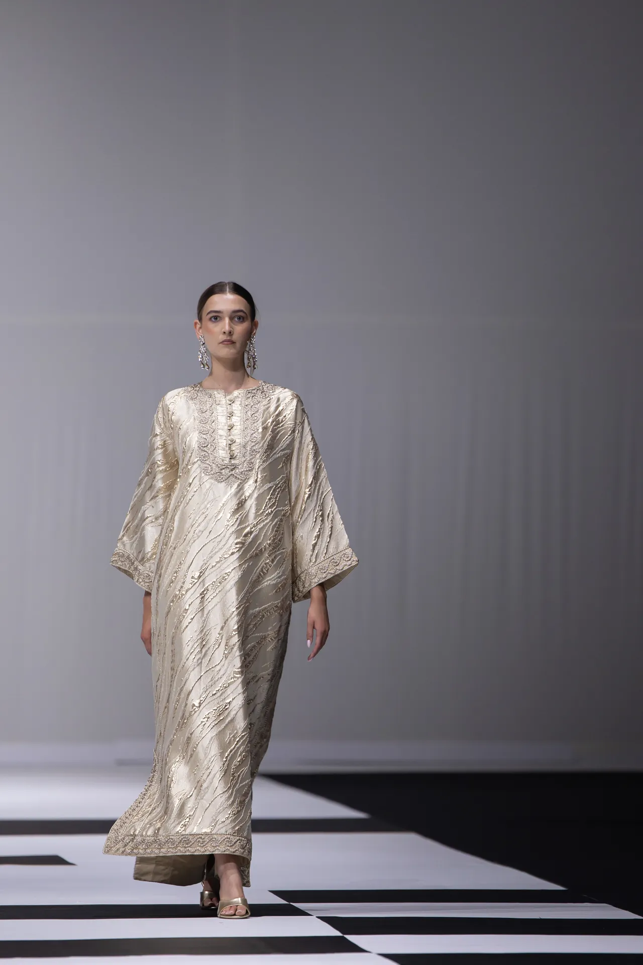 Model walking a fashion runway in a cream-gold embellished gown with big dangling earrings.